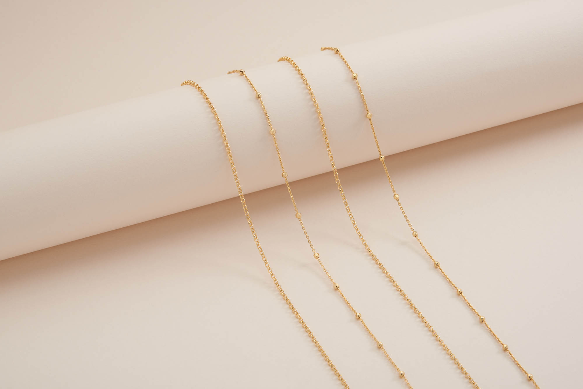 Four delicate gold chain necklaces with small bead accents are draped over a cream-colored cylindrical display on a neutral background.