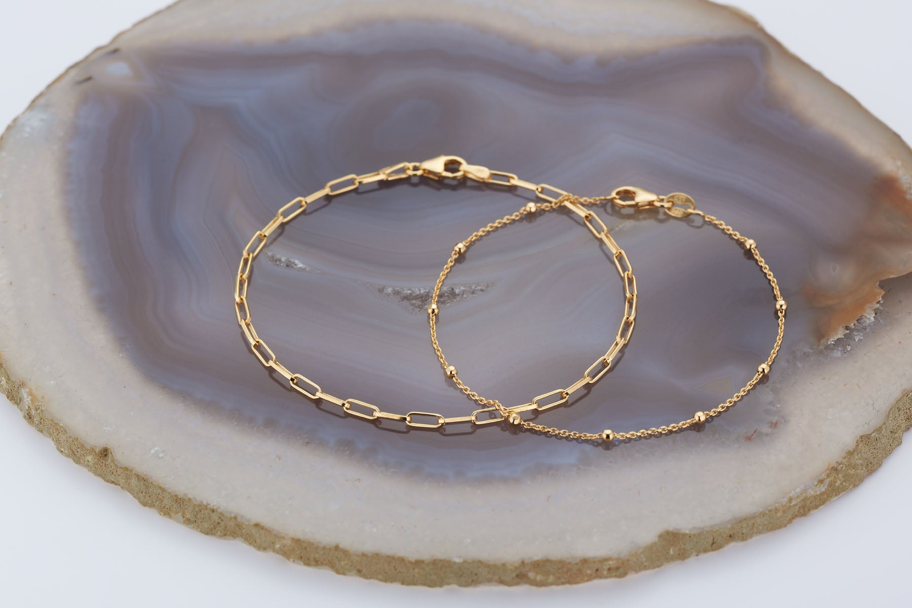 The Baby Paperclip Bracelet and a classic gold chain are showcased atop a polished, circular agate stone slab featuring elegant gray and beige layers.