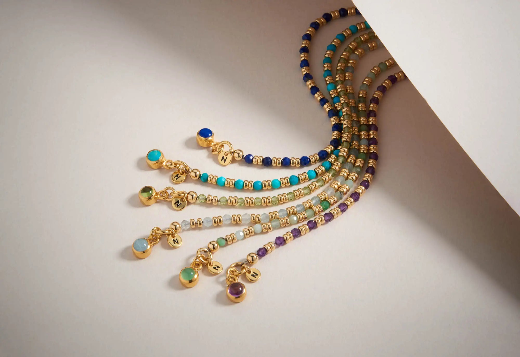 Five beaded bracelets with gold accents and colorful beads—blue, purple, turquoise, and teal—are partially tucked under a white sheet, each ending with a gold charm and a small colored gemstone.