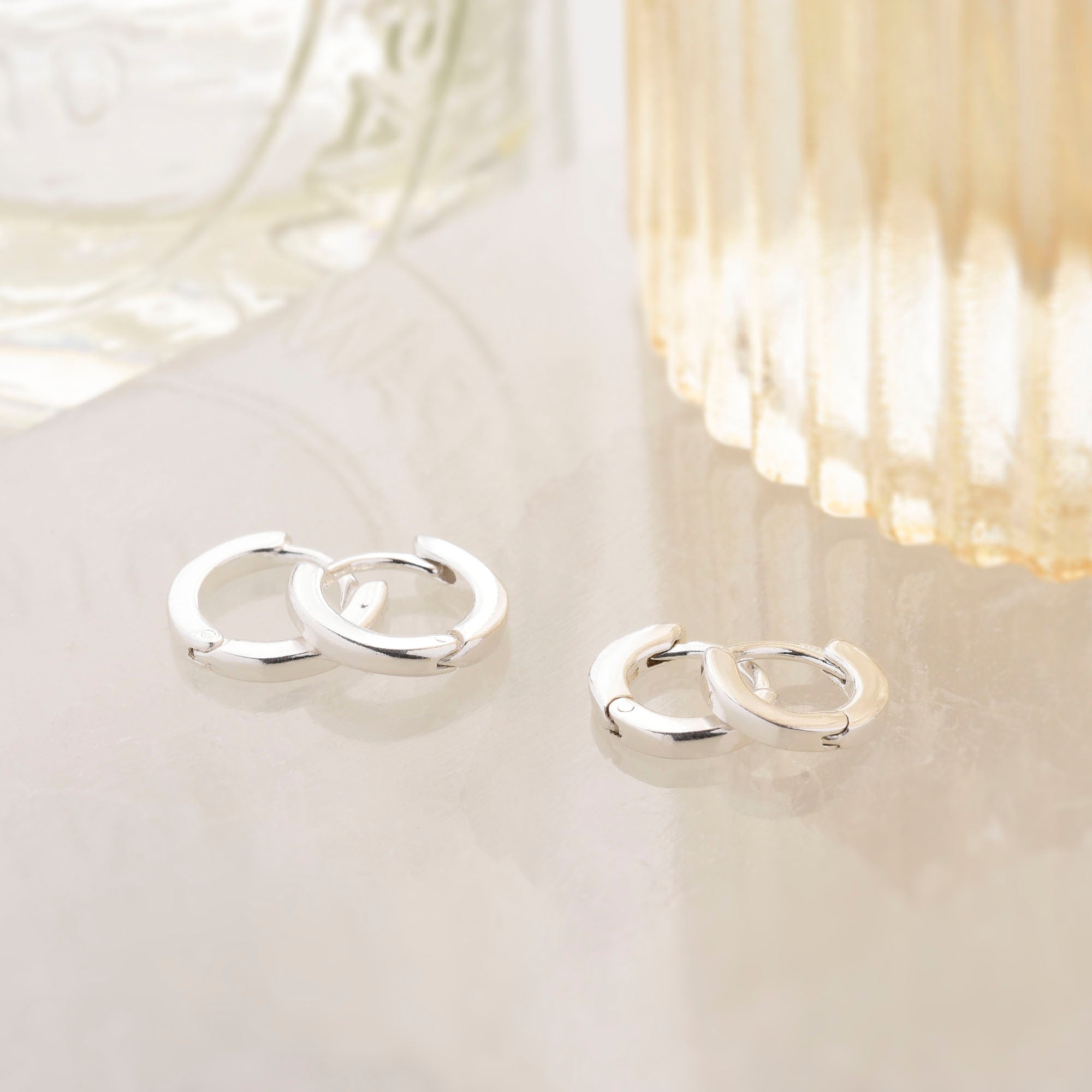 Two pairs of Nell Huggie Earrings in sterling silver, featuring a minimalist style, rest on a light surface near textured glass containers. The larger hoops are positioned on the left, while the smaller ones are on the right.