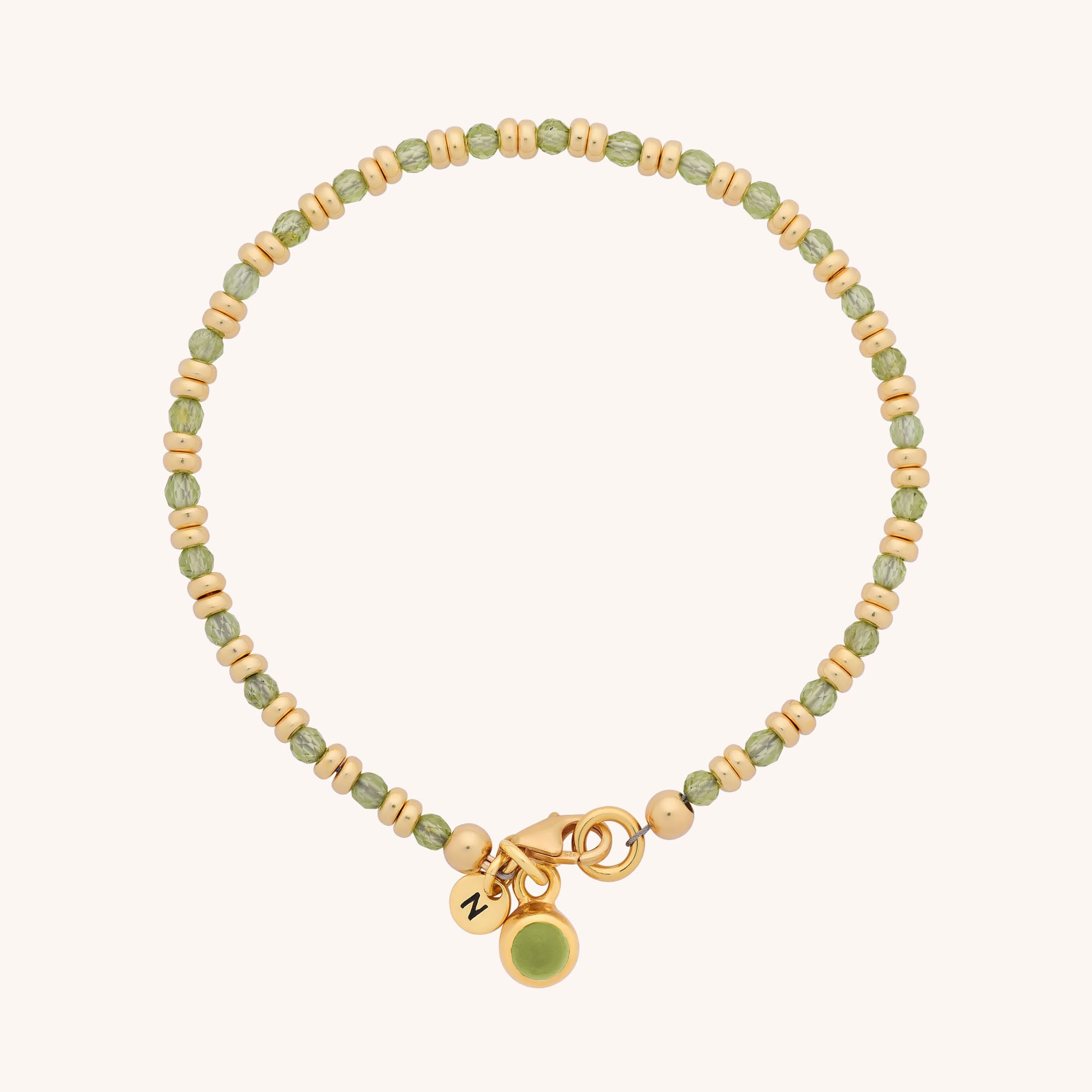 The Linear Birthstone Bracelet features alternating green and gold beads, natural gemstones, a gold clasp, and two dangly gold charms—one engraved with a letter, one with a green stone. Ideal for fans of semi-precious birthstone jewelry.