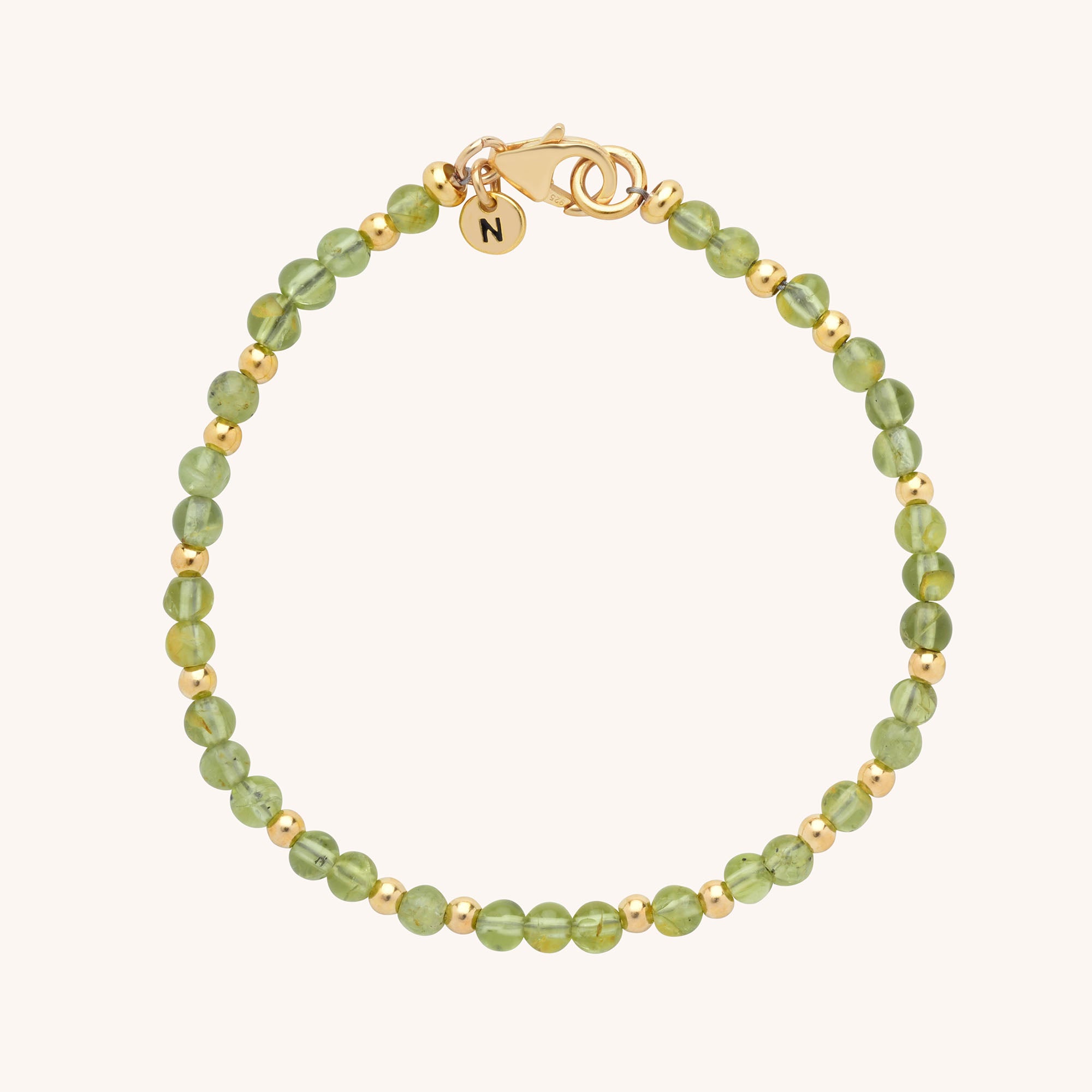 A delicate handmade jewellery piece, the Jewel Bead Birthstone Bracelet features alternating small green and gold beads, a gold clasp, and a round gold charm engraved with the letter N on a light background.