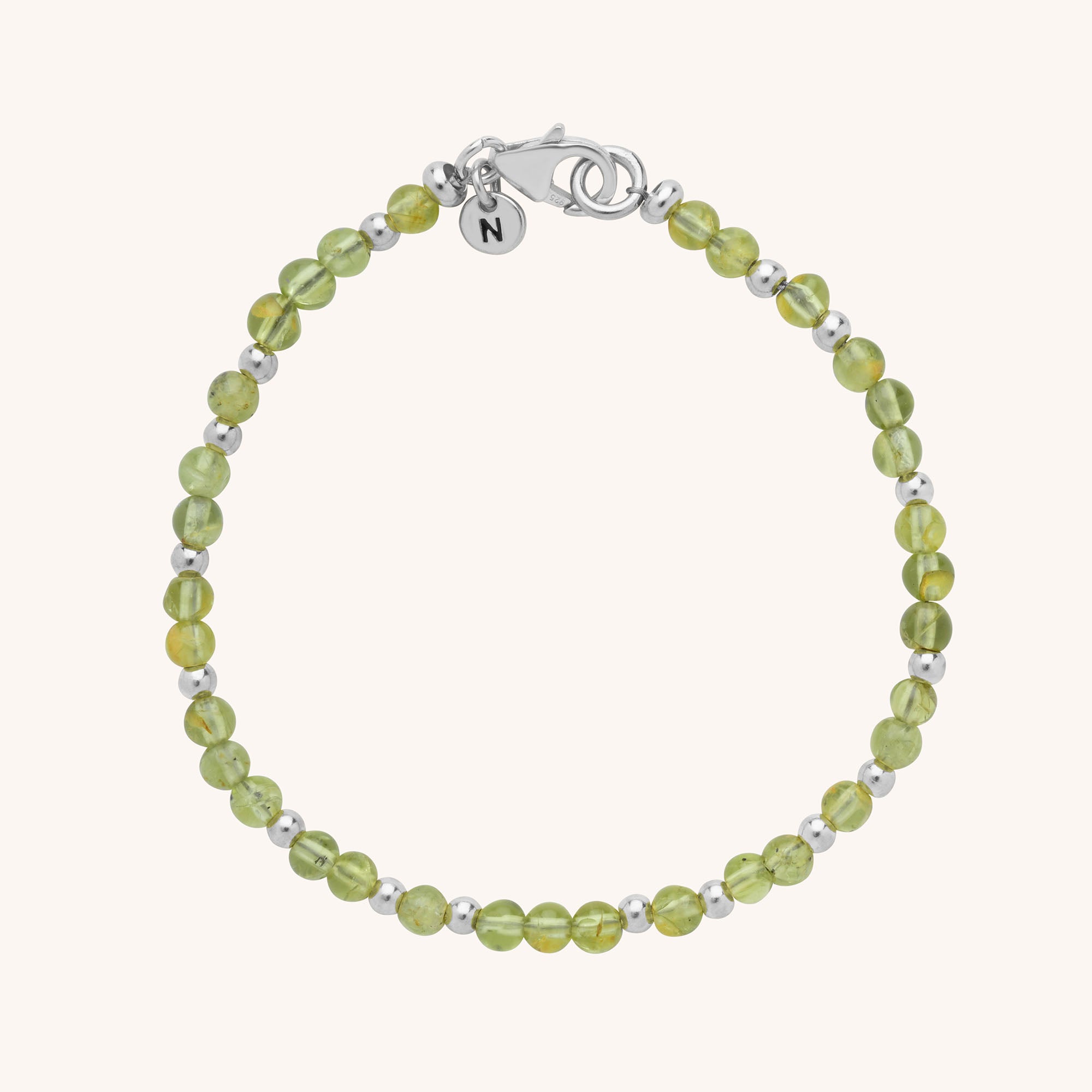 The Jewel Bead Birthstone Bracelet is a delicate handmade piece featuring light green round beads, silver accents, a silver clasp, and a small round charm engraved with the letter N.