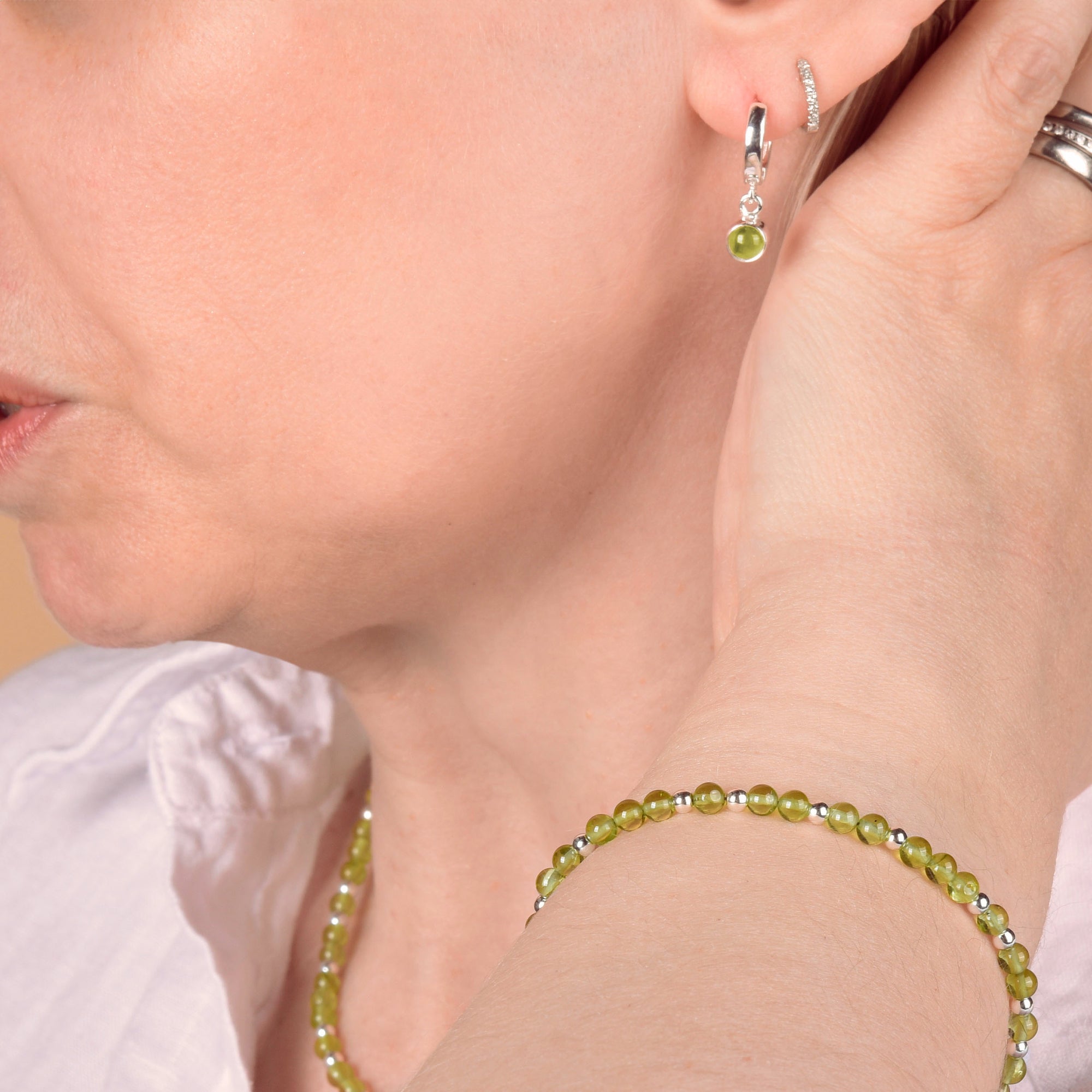 A person in a white shirt displays the Jewel Bead Birthstone Bracelet - August with green peridot beads, a matching peridot necklace, small green earring, silver hoop earring, and silver rings while touching their ear.