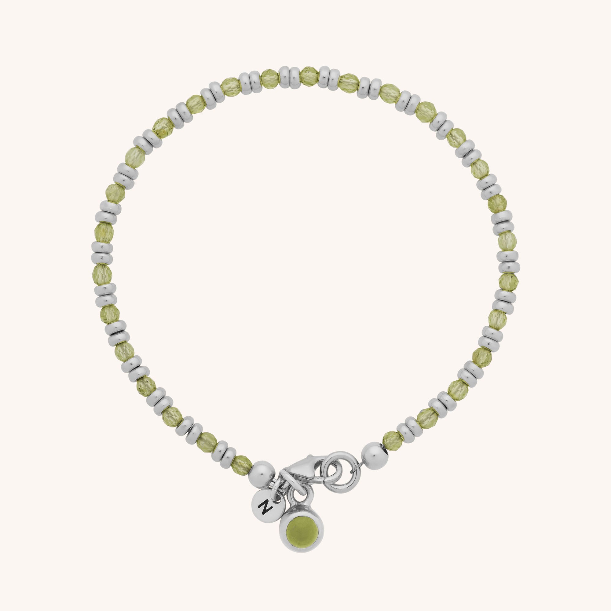 The Linear Birthstone Bracelet is a delicate piece with alternating silver and light green beads, a silver clasp, and two charms—one engraved with “N” and one featuring a green center—ideal for gemstone jewelry lovers.
