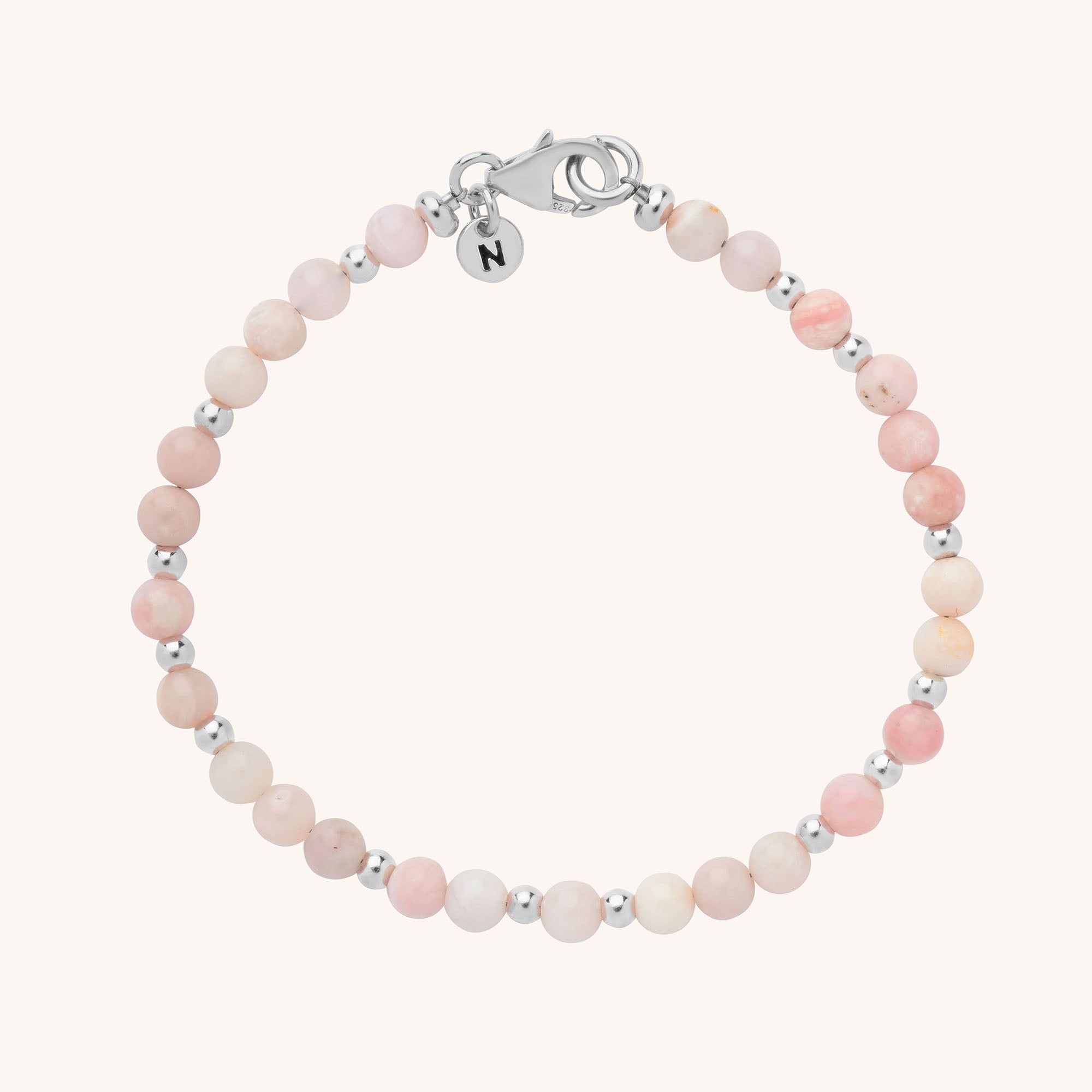 The Jewel Bead Birthstone Bracelet features alternating small round pink and silver beads, a silver clasp, and a round charm engraved with the letter N. This handmade piece is shown on a plain light background.