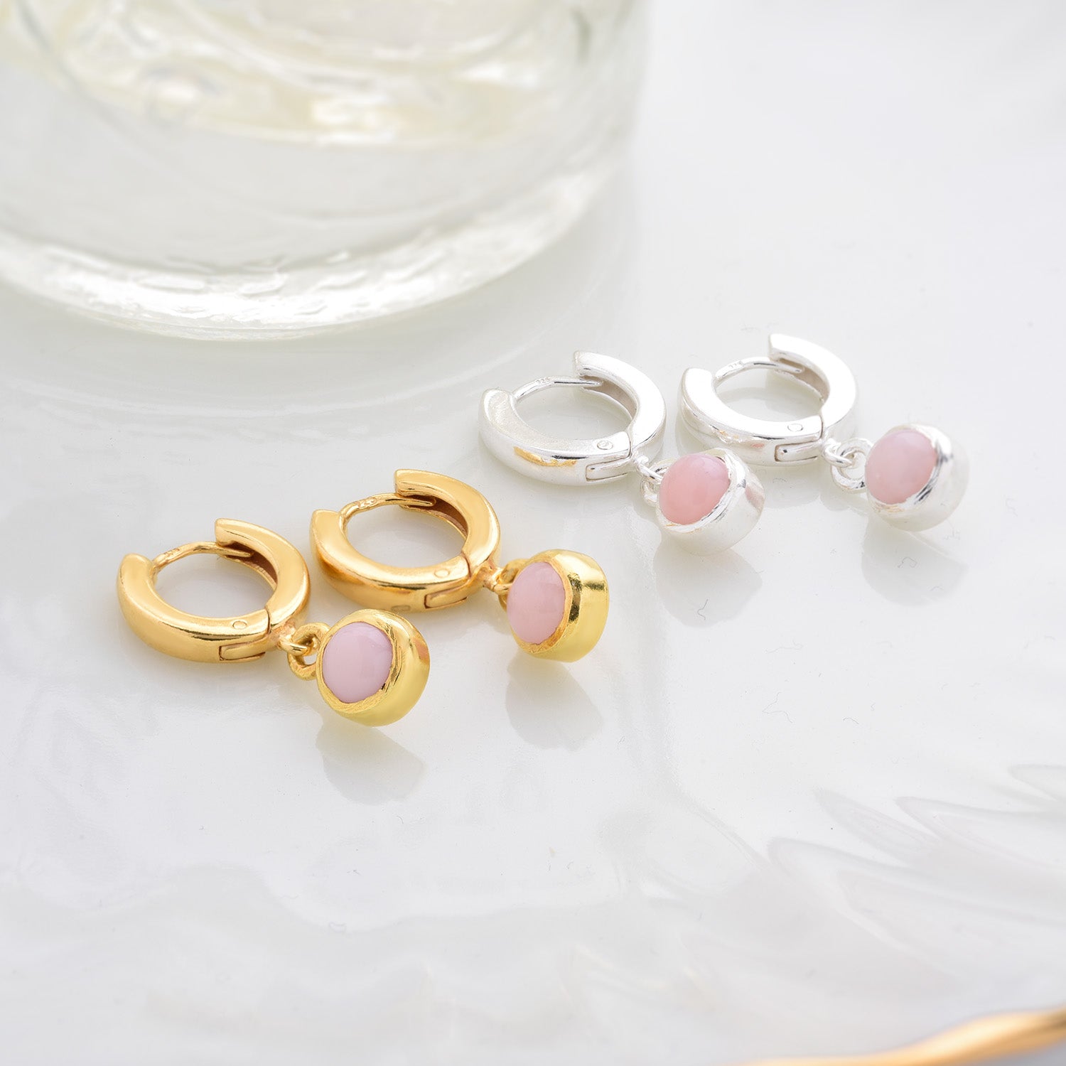 The Gem Hoop Earrings - October Birthstone are shown in gold and silver, each with Pink Opal stones on a white surface near a glass object. Their polished finish enhances their charm, making them perfect for emotional healing.