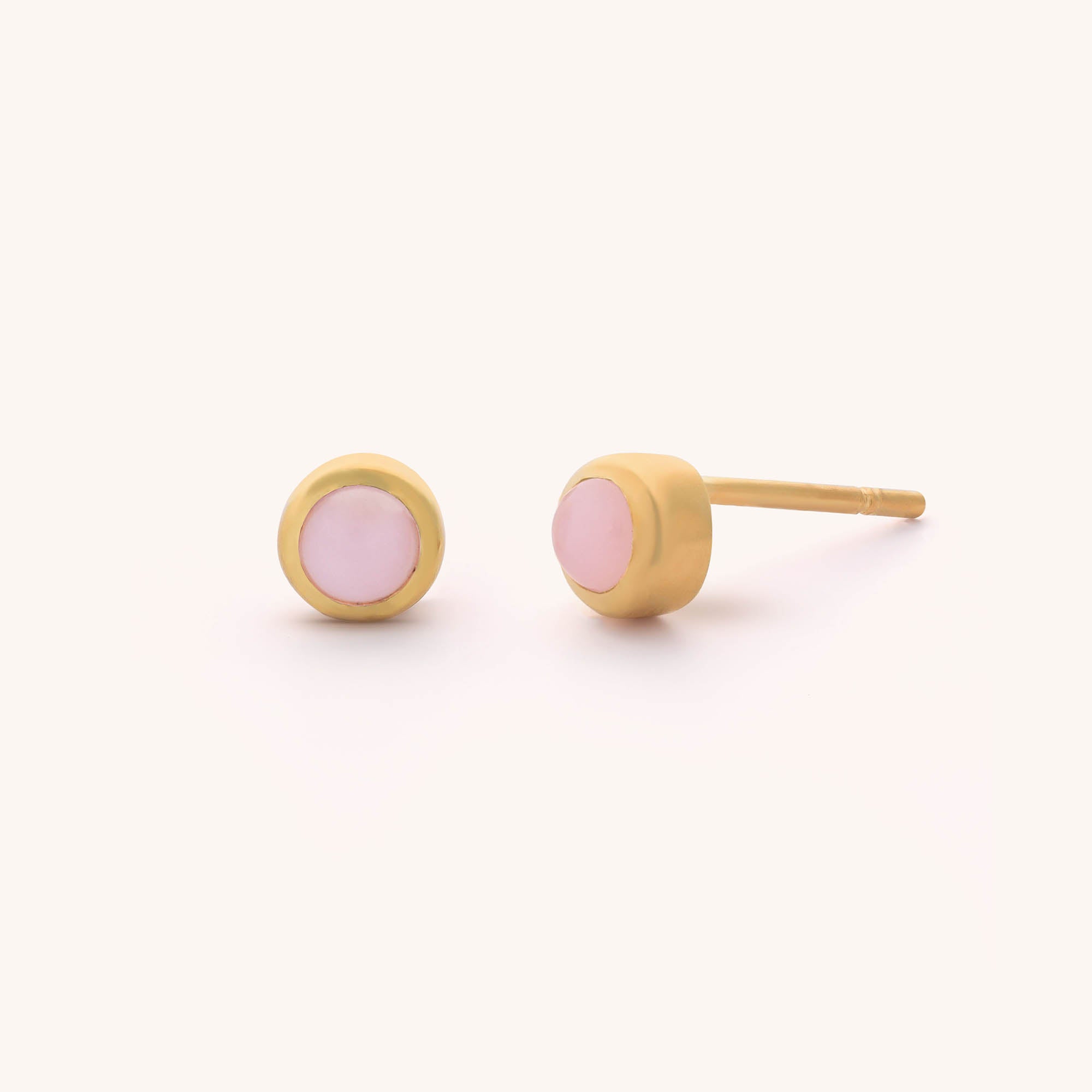 The Mini Gem Dot Birthstone Earrings - October feature a gold pair with round pink opal stone centers, perfect for those born in October. One earring lays flat and the other tilts to show its post, elegantly presented on a clean white backdrop.