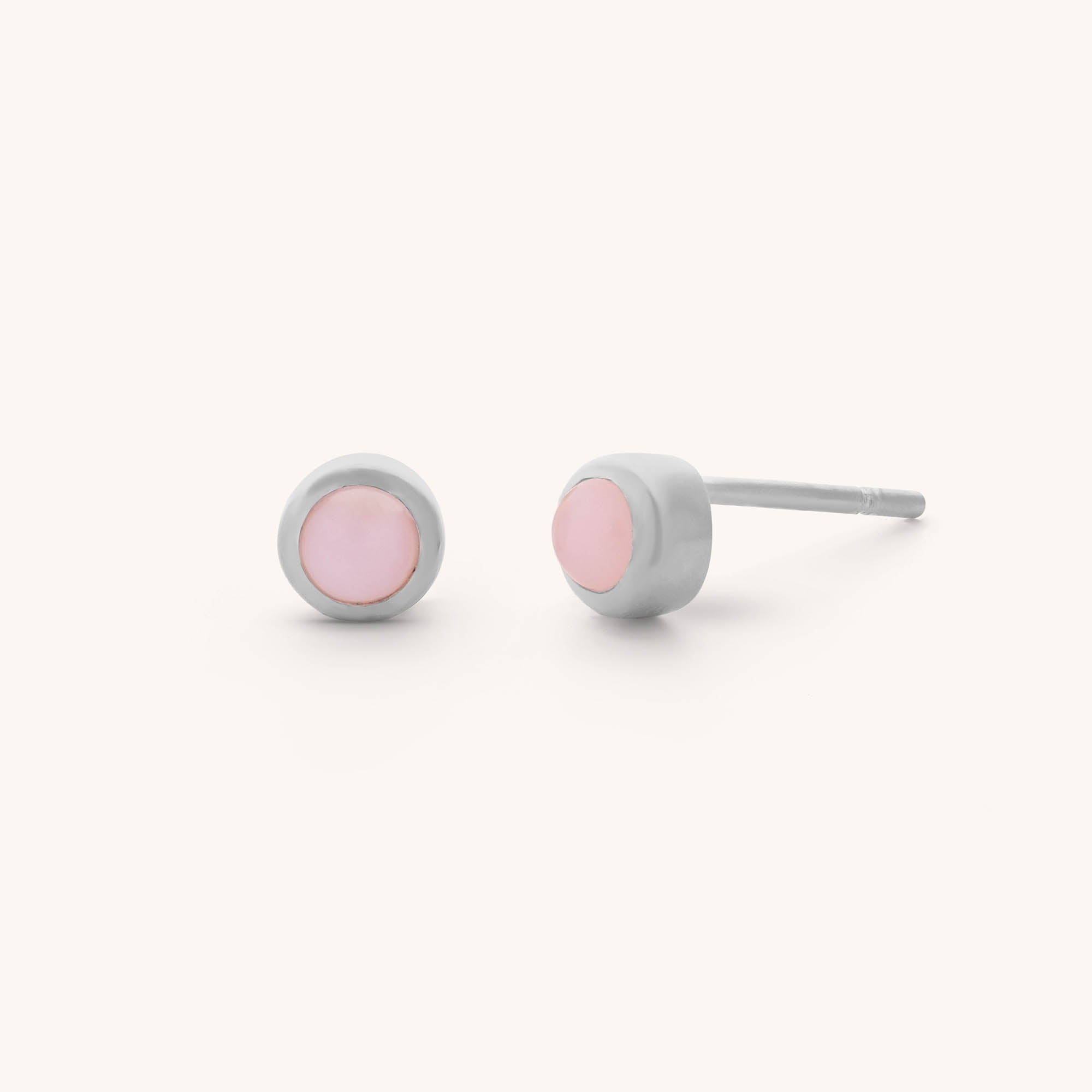 The Mini Gem Dot Birthstone Earrings - October feature circular pink opals set in silver-toned metal. Ideal for October birthdays, these minimalist studs offer a modern and elegant design, beautifully displayed on a light background.