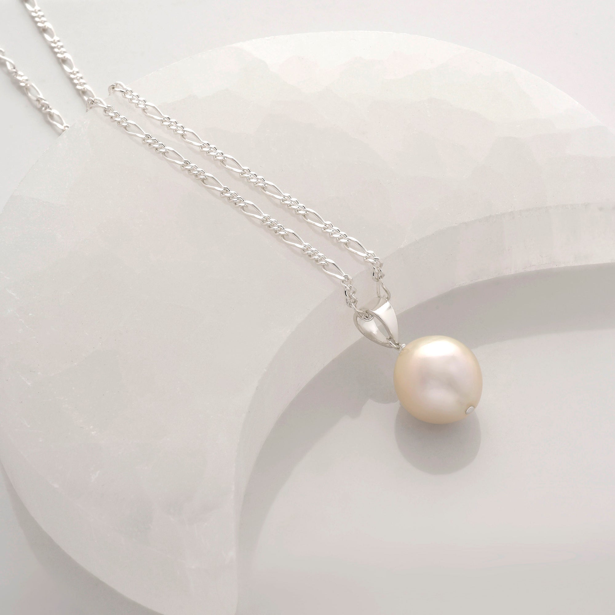 The Ellen Large Pearl Necklace, featuring a single round cultured freshwater pearl on a silver chain, is showcased on a textured white crescent backdrop for a soft, elegant, and minimalist look.