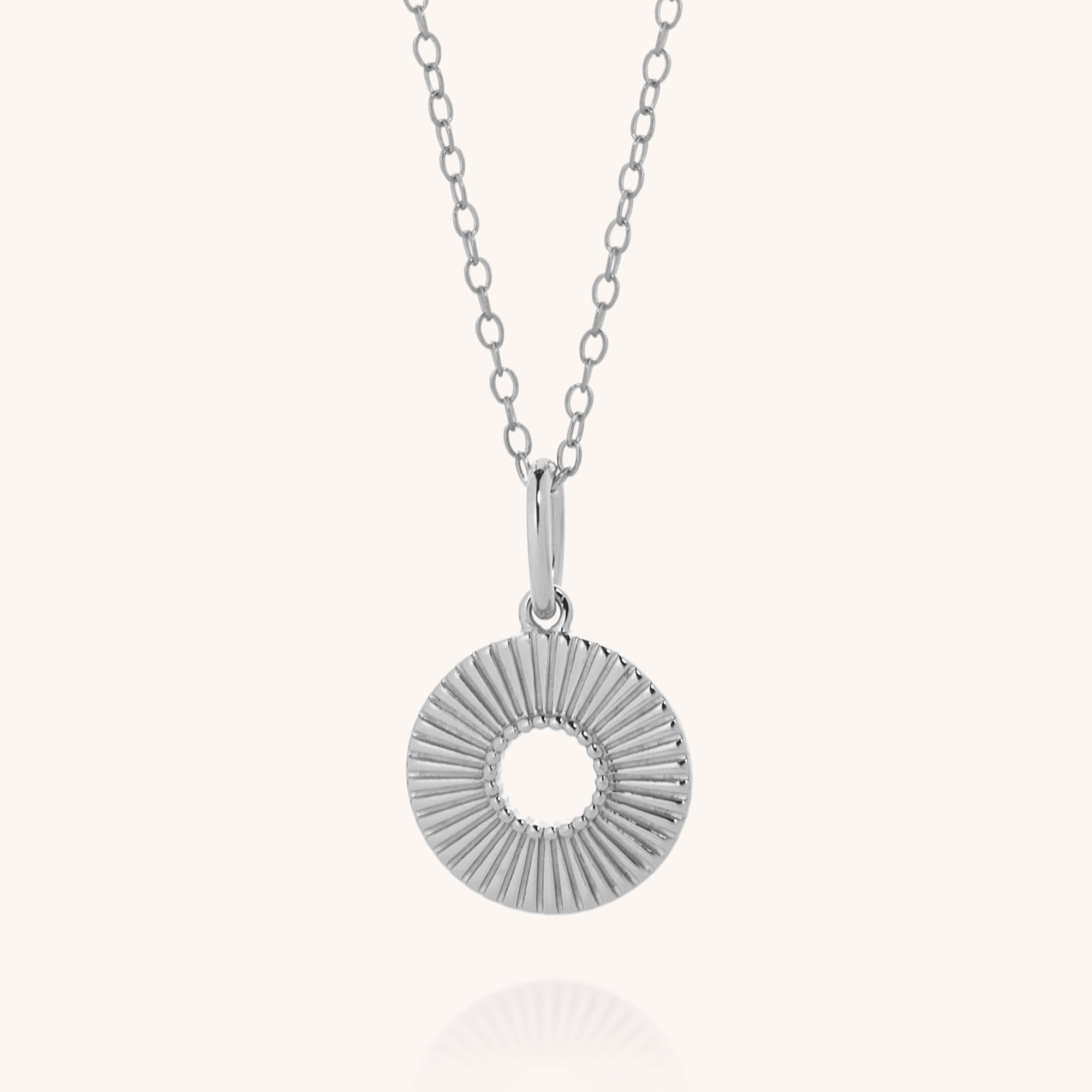 The Solar Circle Necklace features a round, textured sterling silver sunburst pendant with an open center, hanging on a delicate chain against a light background.
