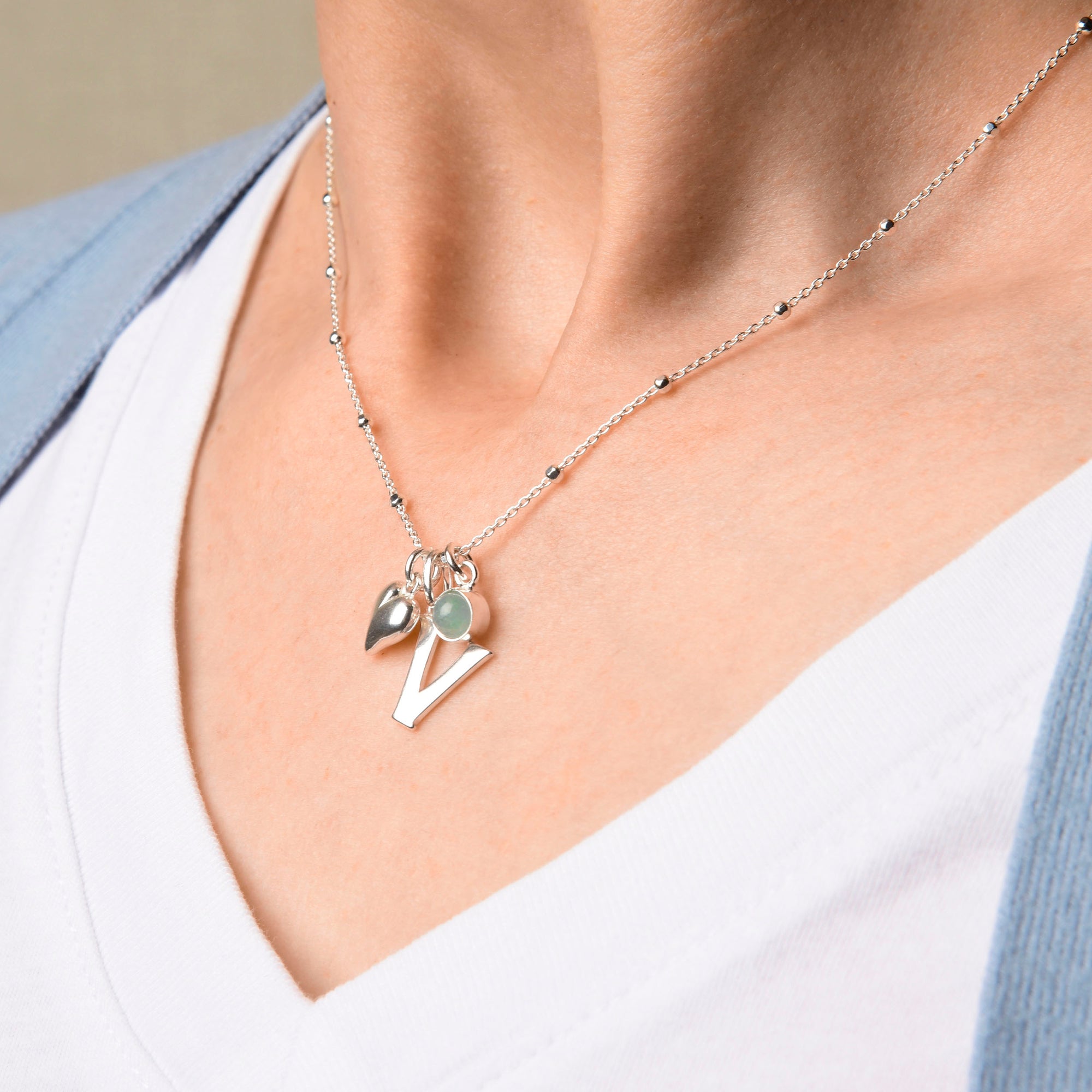 A person wearing the Satellite Chain—an eco sterling silver necklace with a heart charm, round gemstone, and letter V—over a white shirt and light blue cardigan.