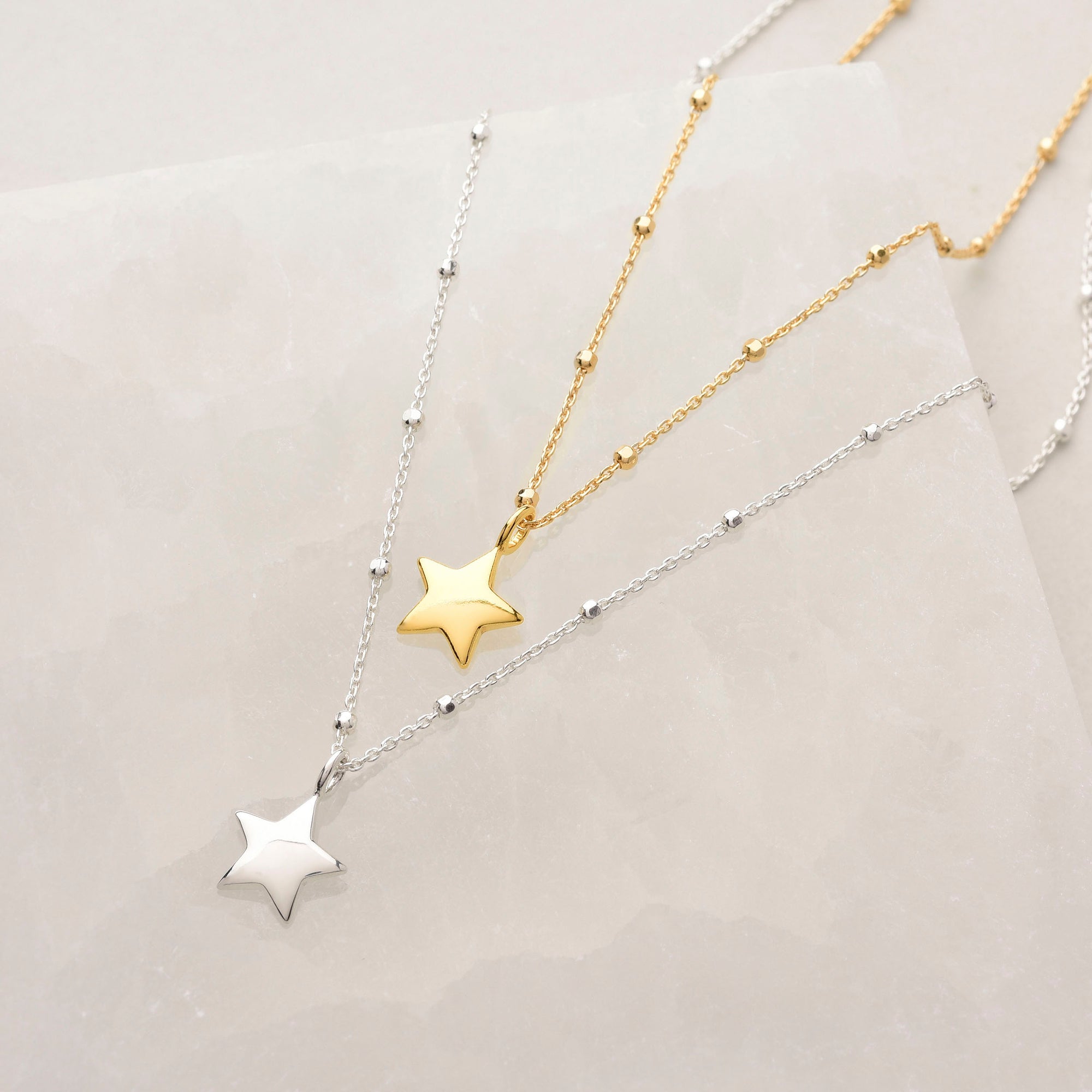 Two Star Charm Necklaces—one in eco-friendly silver and one in gold—rest on a pale, textured surface. Each features bead accents along the chain, with the star pendants displayed side by side from our celestial-inspired collection.