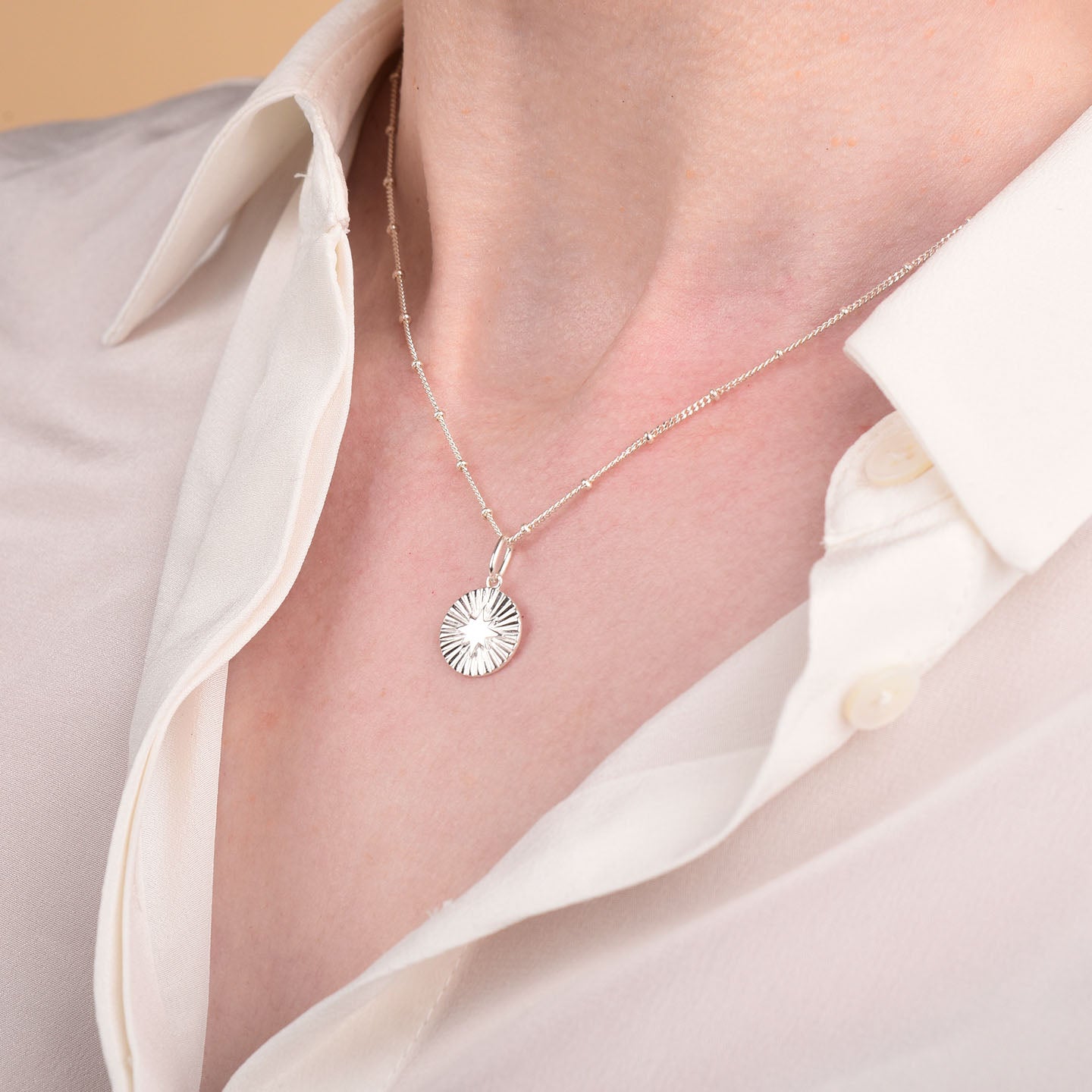 A close-up of a person wearing a white button-up shirt and the Shining Star Necklace, which features a round, sunburst pendant for celestial flair.