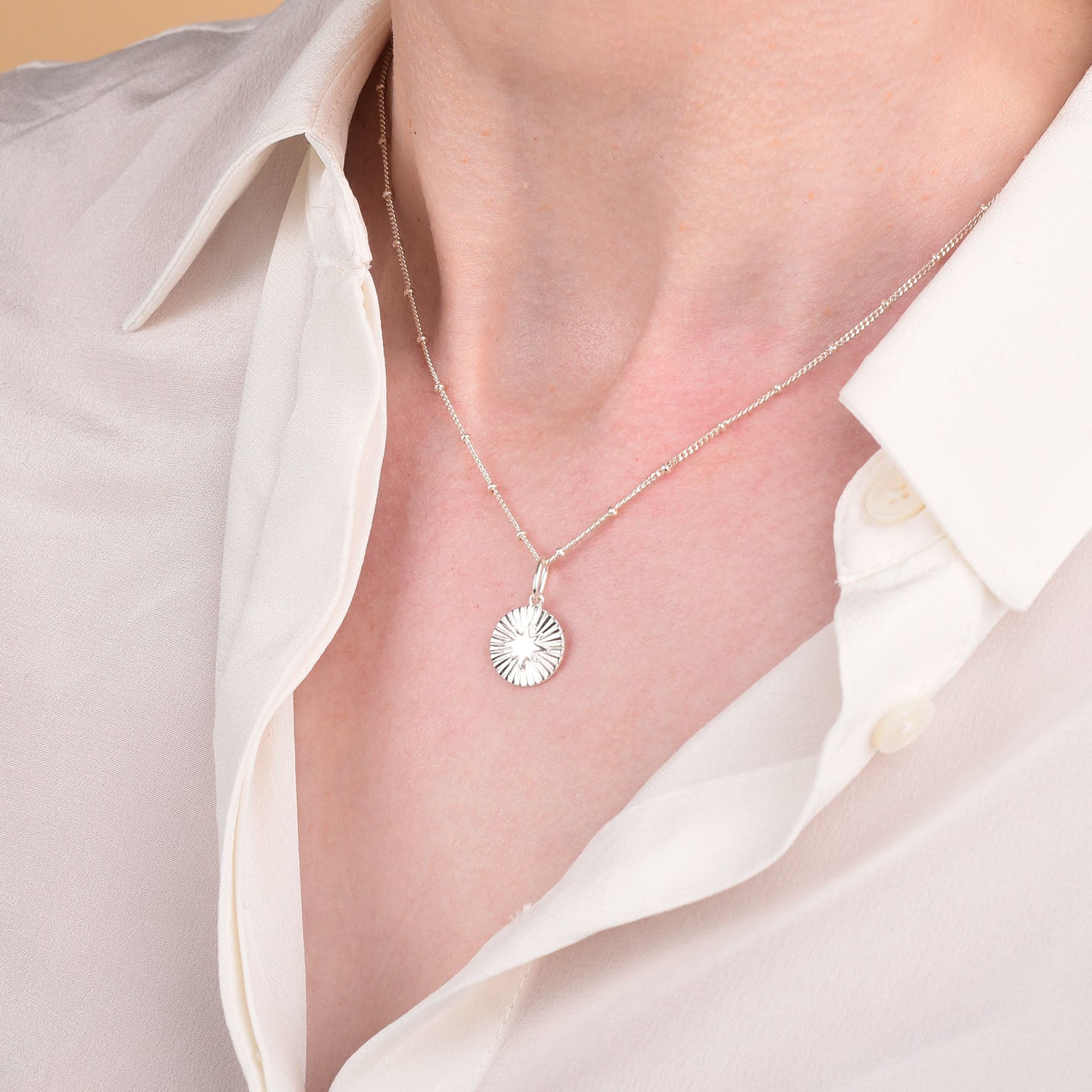 Wearing a white collared shirt, slightly open at the neck, a person highlights the Shining Star Necklace—a sterling silver piece with a round textured pendant—emphasizing its celestial-inspired style.