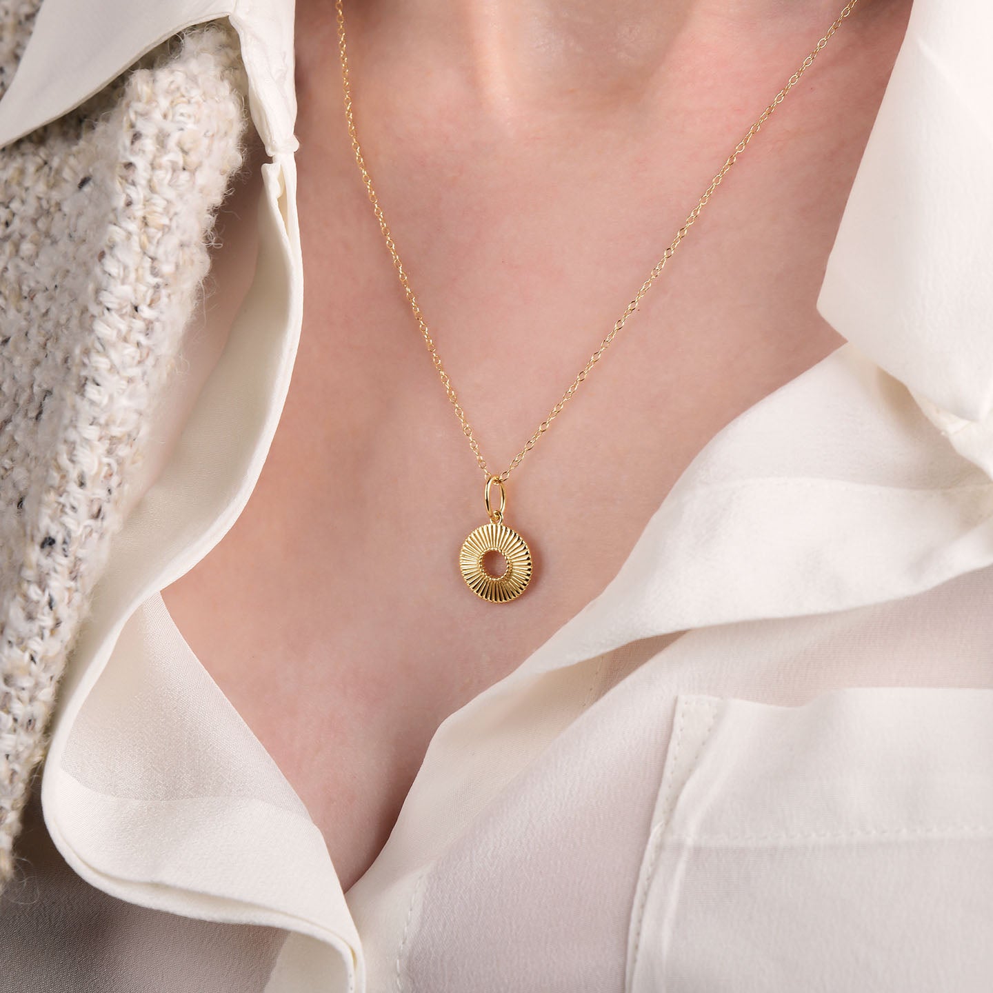 A person in a textured cream jacket and white blouse wears the Solar Circle Necklace, featuring a gold-plated sterling silver pendant with an eye-catching sunburst design.