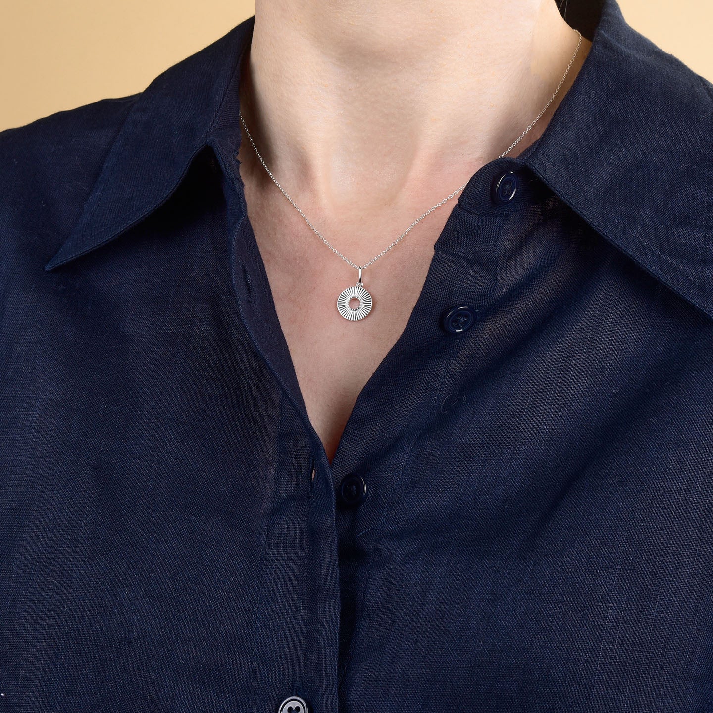 A person wears the Solar Circle Necklace in sterling silver, featuring a circular pendant, along with a dark navy blue button-up shirt, shown from shoulders to upper chest against a beige background.