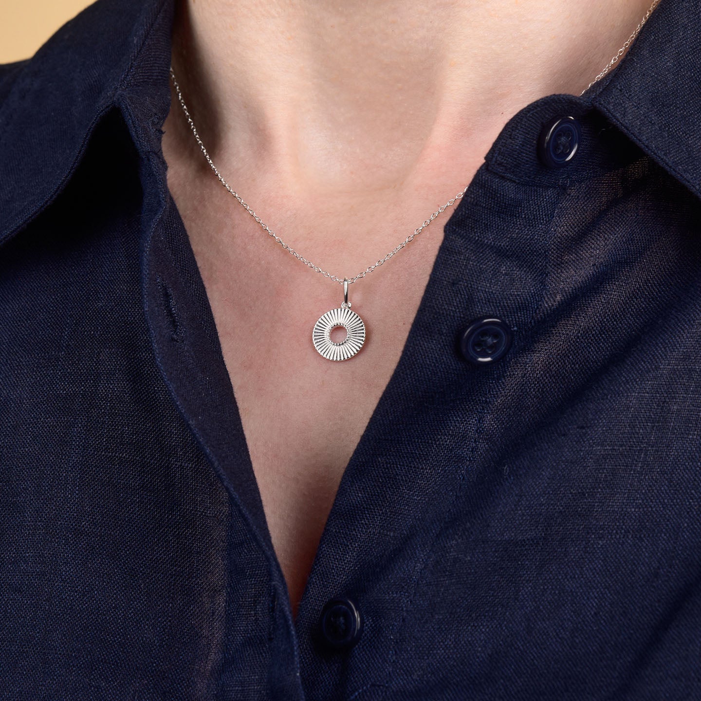 A person wearing a navy blue button-up shirt and the Solar Circle Necklace, which features a round sunburst-patterned pendant.
