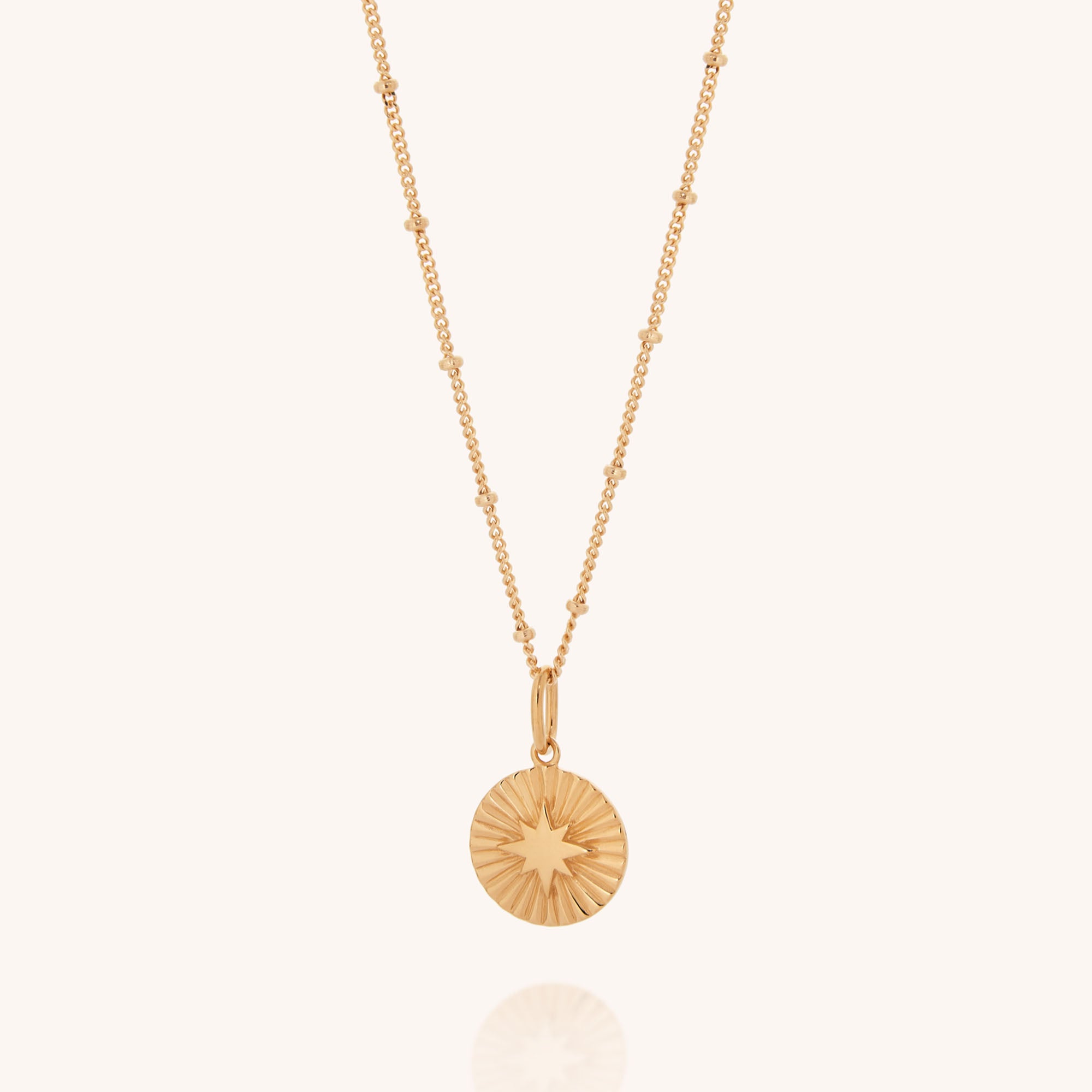 The Shining Star Necklace features a delicate gold beaded chain and round pendant with a radiant sunburst design, offering celestial style beautifully presented against a light background.