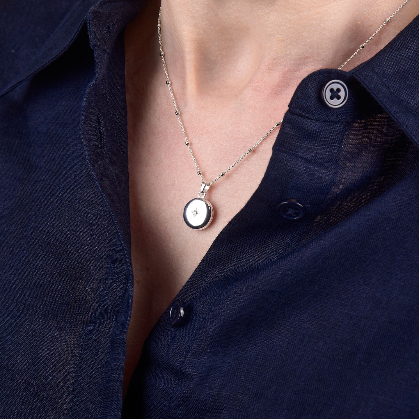 A person wearing a navy blue button-up shirt and the Starlight Keepsake Locket Necklace, a sterling silver celestial-themed locket, photographed close-up at the neck and collar.