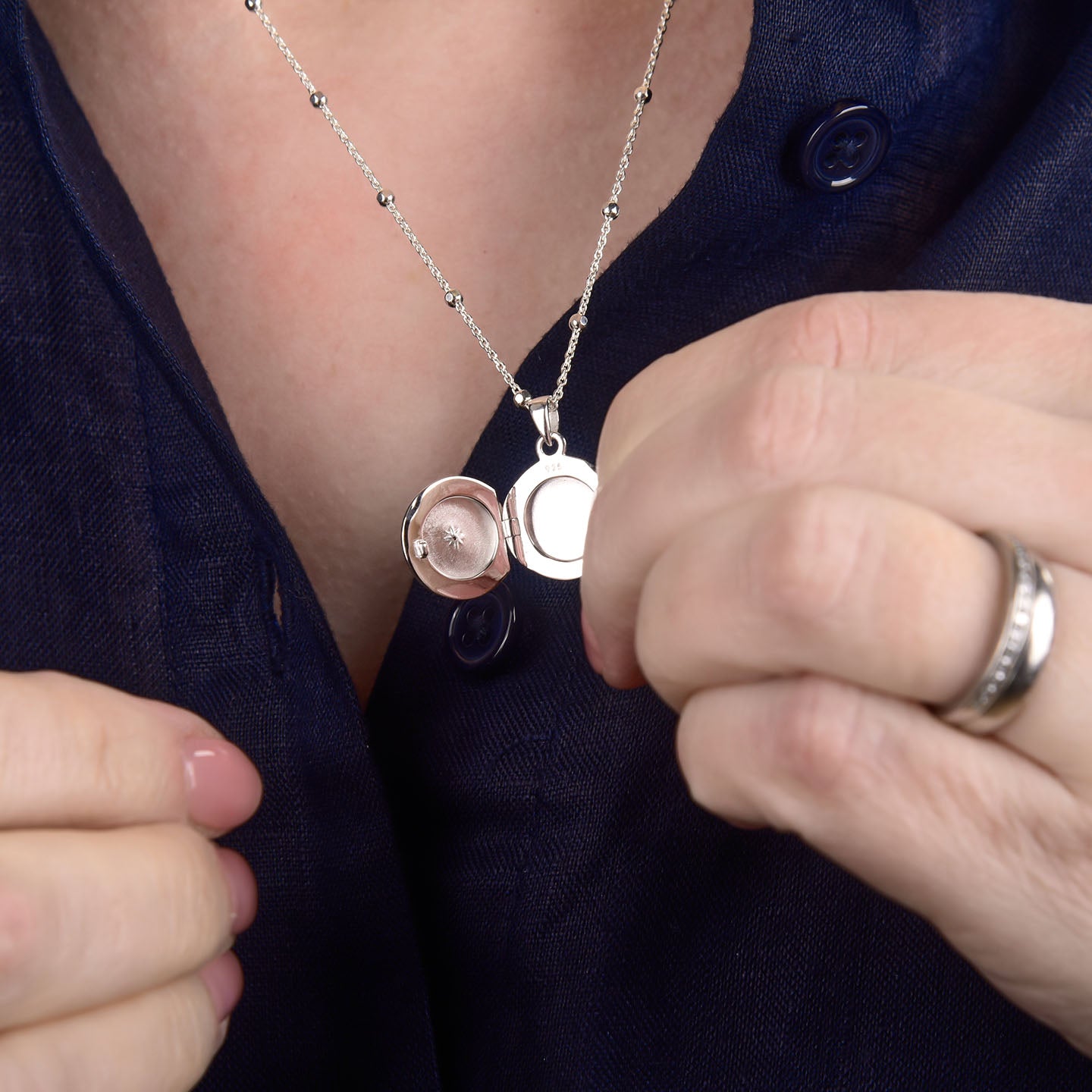 A person wearing a dark blue shirt displays the elegant Starlight Keepsake Locket Necklace, holding it open to reveal its empty interior. Their pink nail polish and ring accentuate the sterling silver locket’s timeless style.