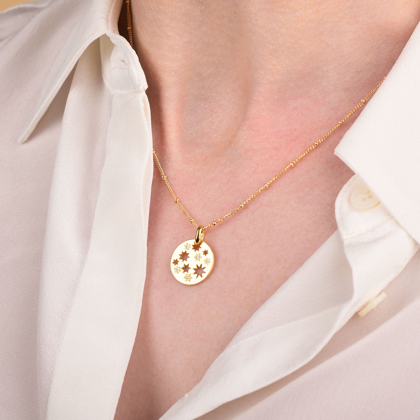 A person wearing a cream-colored blouse and the Starry Skies Necklace—a gold necklace featuring a round pendant with star-shaped engravings and subtle cubic zirconia accents.