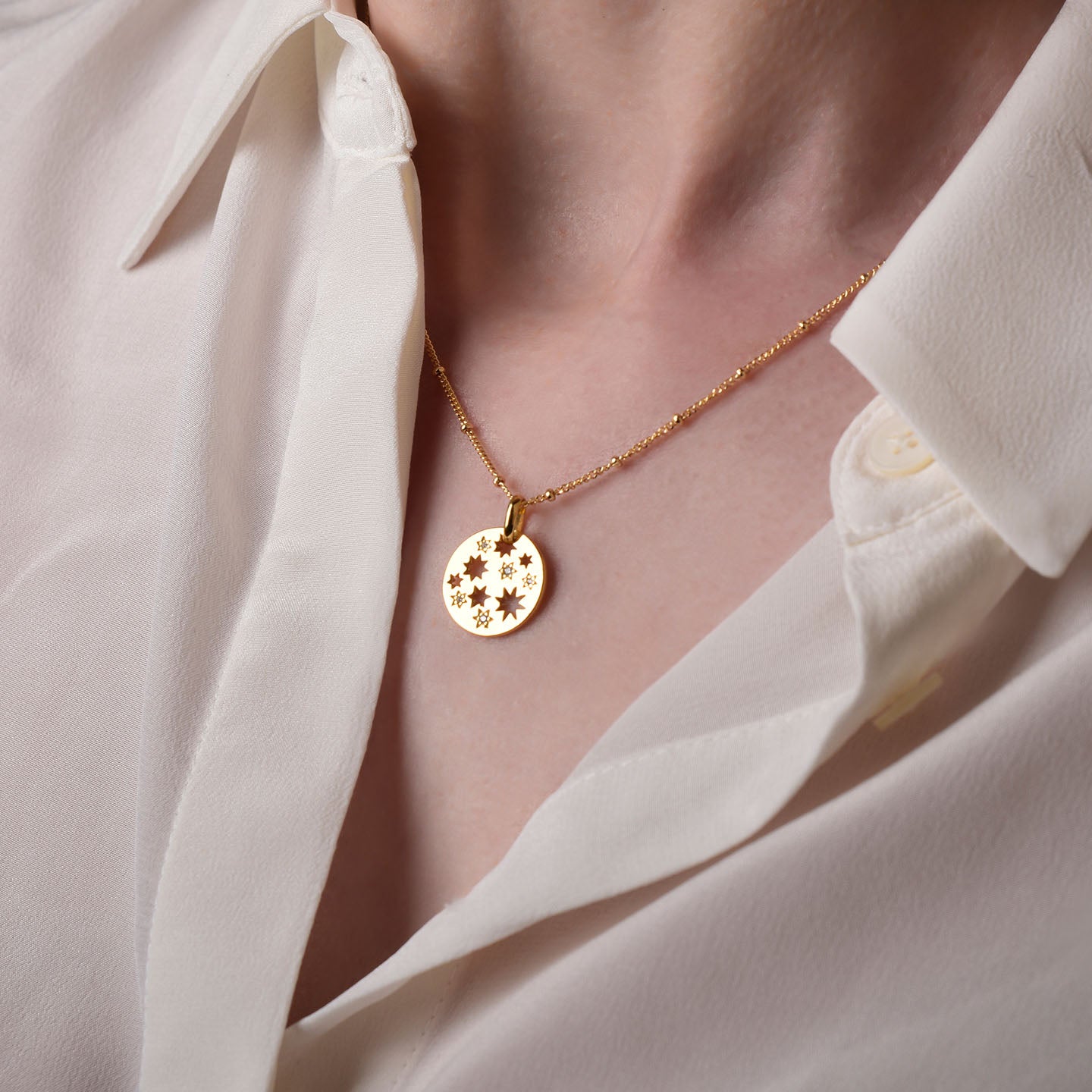 A close-up of a person wearing a white button-up shirt and the Starry Skies Necklace, featuring a gold round pendant engraved with stars and cubic zirconia accents.