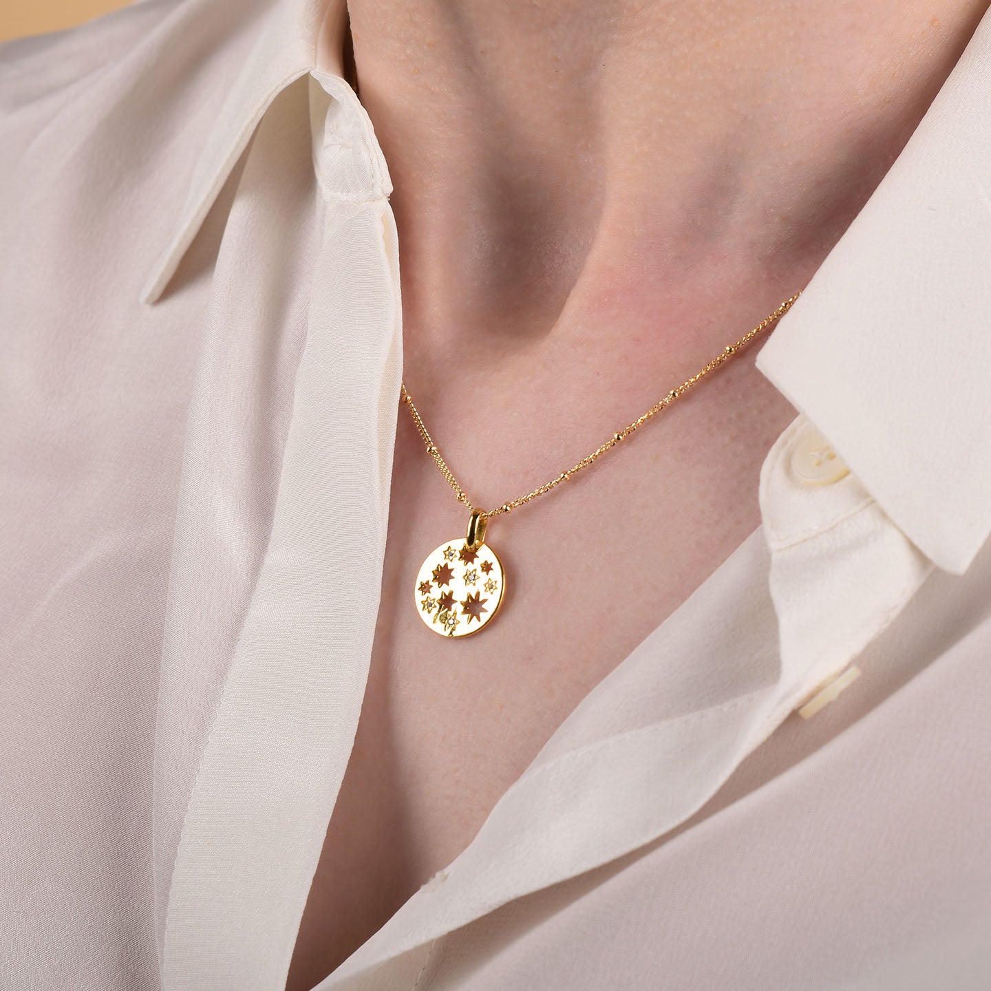 A person wears an open-collared white shirt and the Starry Skies Necklace, a gold celestial piece featuring a round pendant with delicate flower engravings and sparkling cubic zirconia accents.