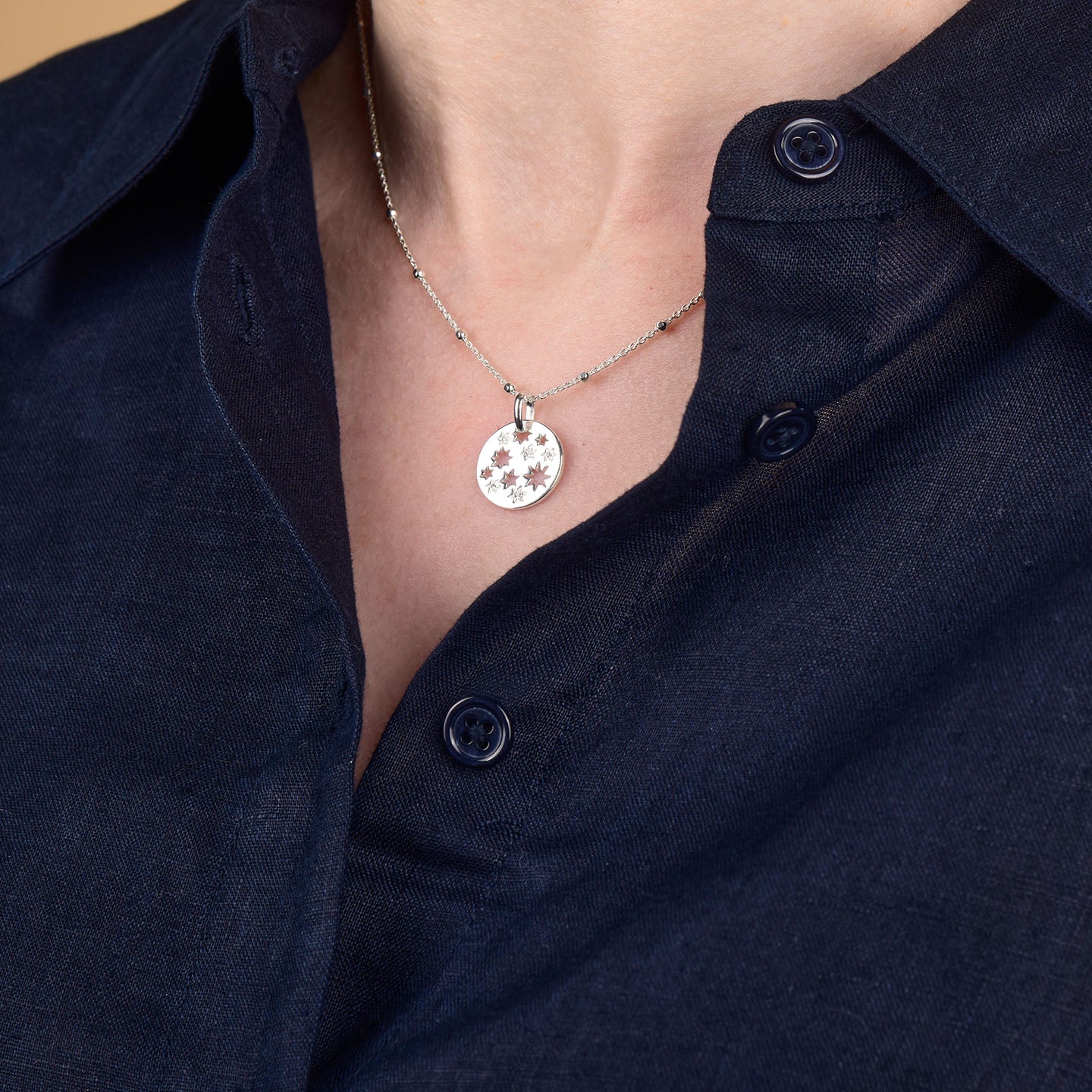 A close-up of a person wearing a dark navy blue button-up shirt and the Starry Skies Necklace, which features a silver round pendant adorned with a starburst design and sparkling cubic zirconia accents.