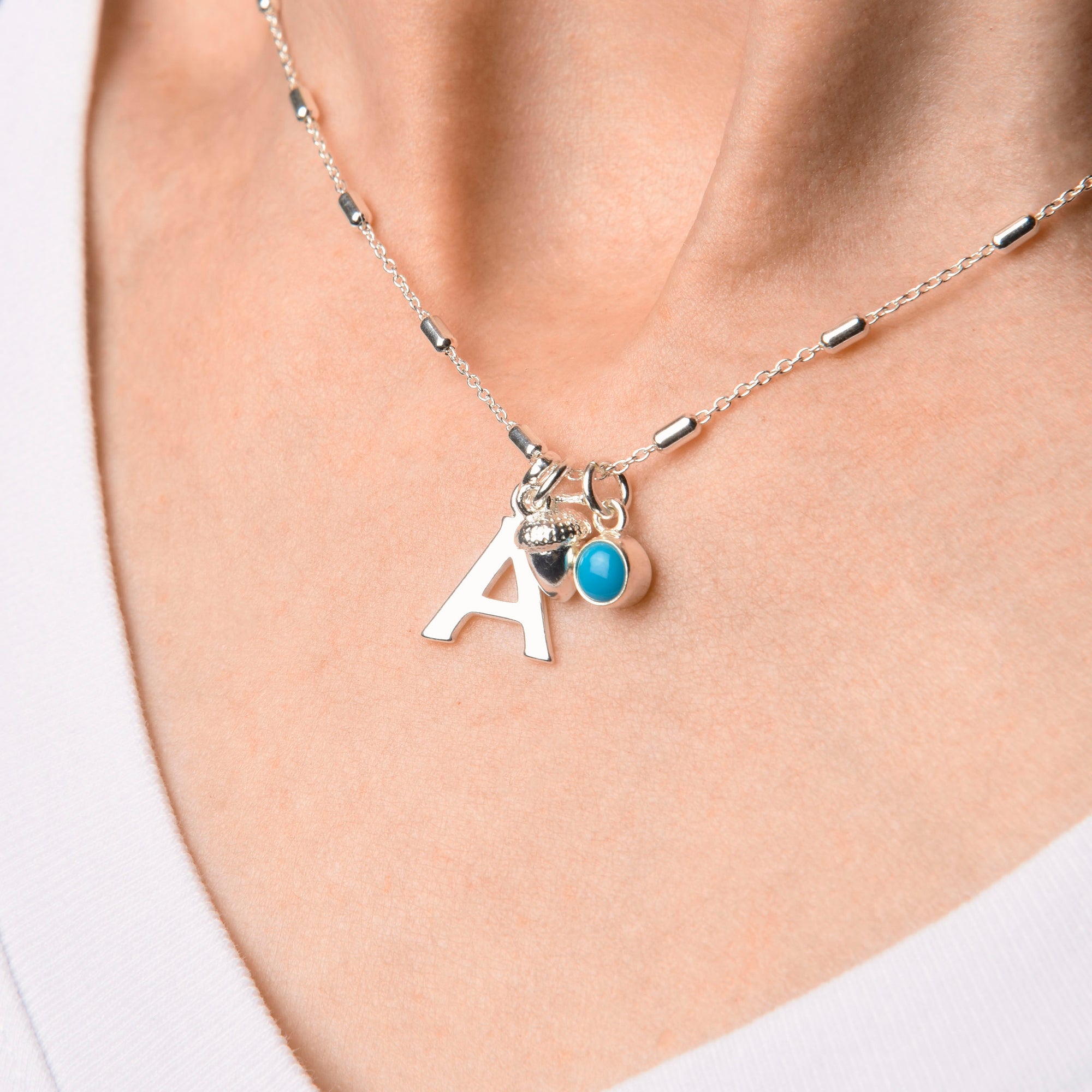 A close-up of the Tubular Satellite Chain, a recycled sterling silver necklace with a letter A pendant, textured charm, and round blue stone, styled with a white V-neck shirt.