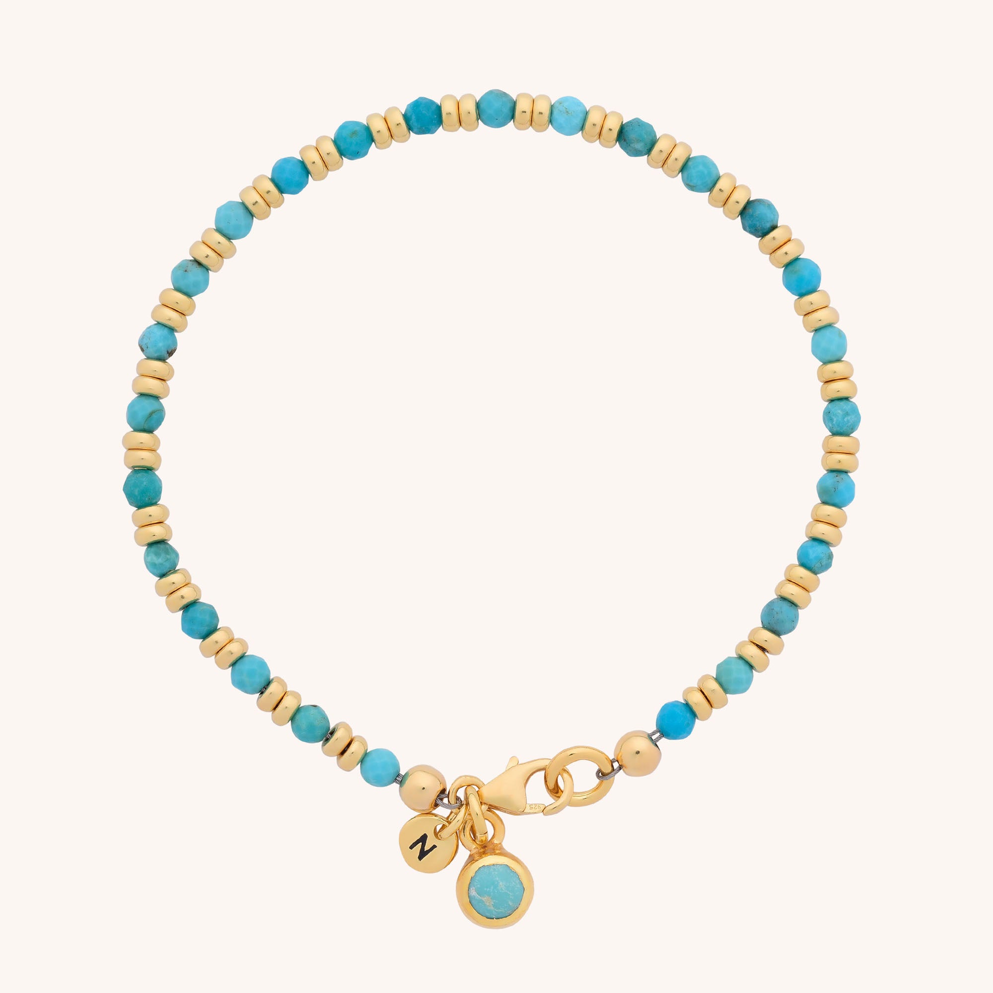 The Linear Birthstone Bracelet features delicate turquoise and gold beads, a gold clasp, and two dangling charms: an engraved gold disc and a turquoise gemstone set in gold.