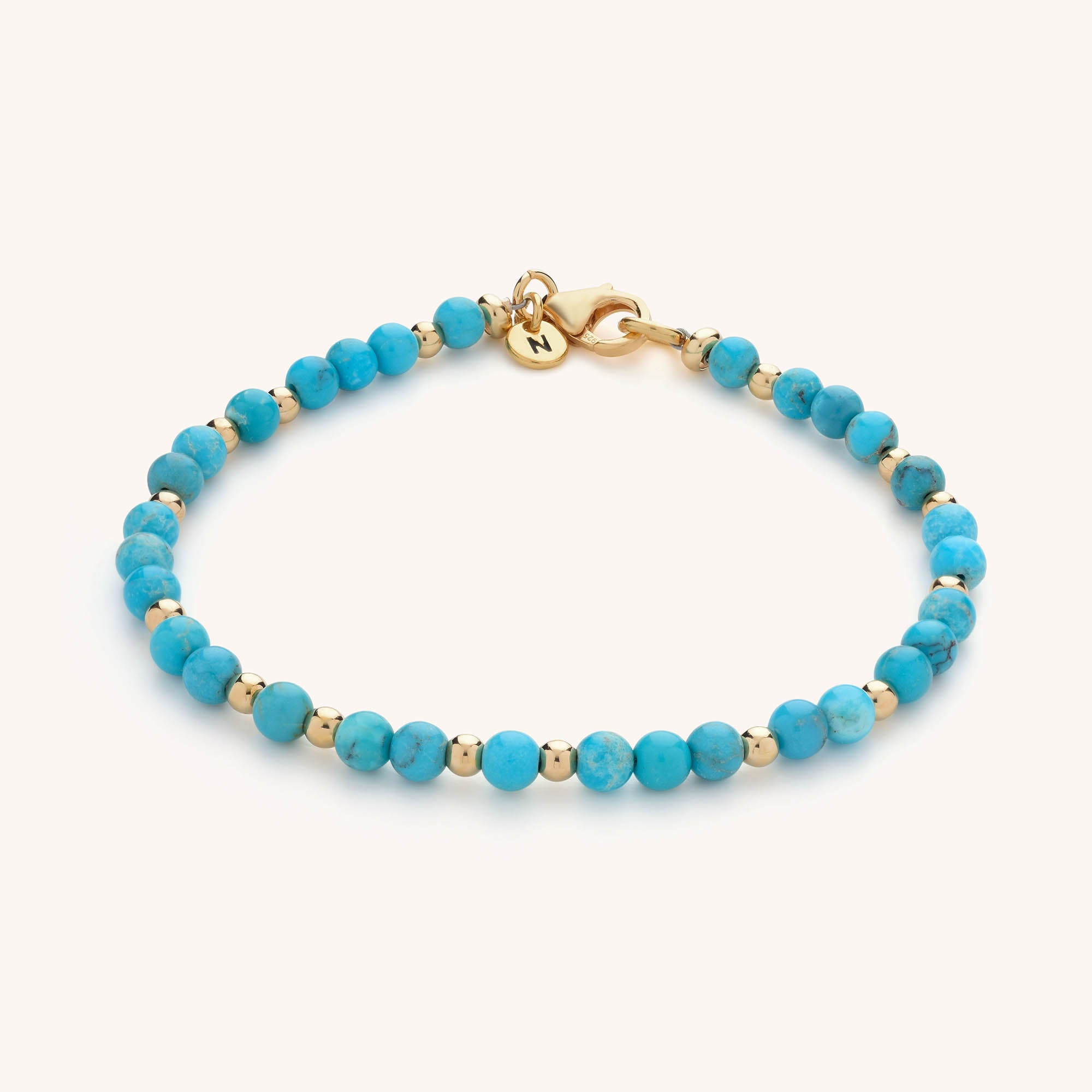 The Jewel Bead Birthstone Bracelet features round turquoise and gold beads, a gold clasp, and a small round N-engraved charm—an elegant piece of handmade jewellery.