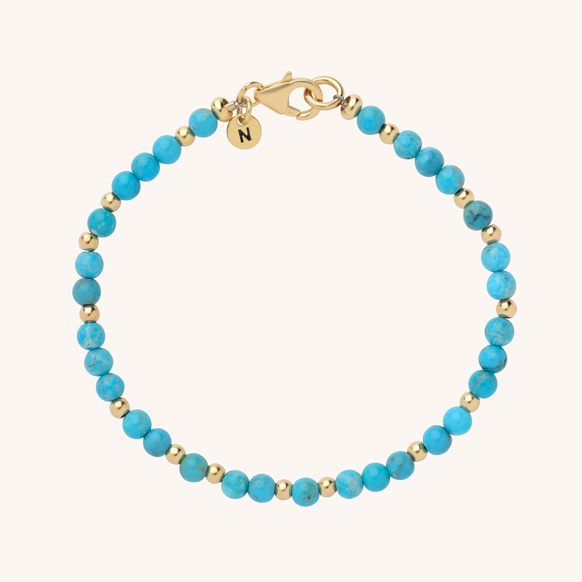 The Jewel Bead Birthstone Bracelet is a handmade piece with alternating small turquoise and gold beads, finished with a gold clasp and a round gold charm engraved with the letter N.