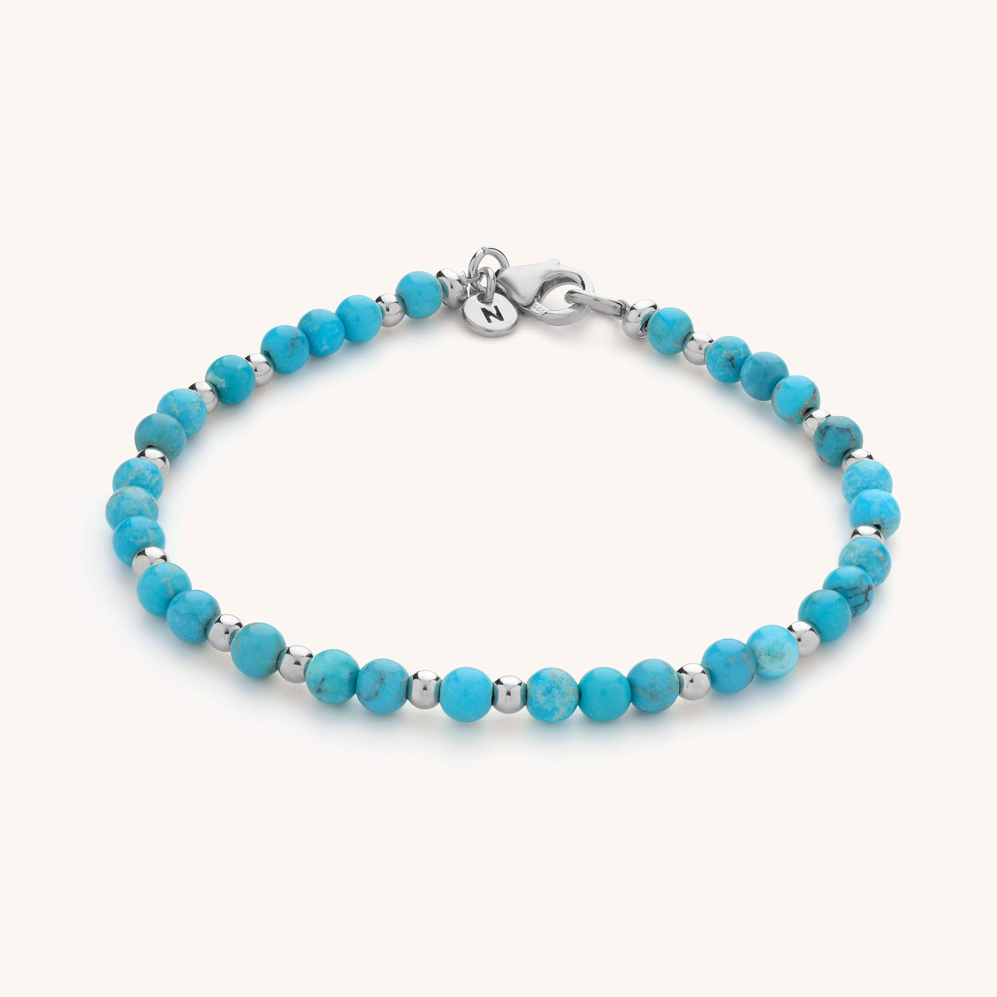 The Jewel Bead Birthstone Bracelet is a delicate handmade piece featuring small round turquoise beads, metallic silver spacers, a silver clasp, and a round tag set against a light background.