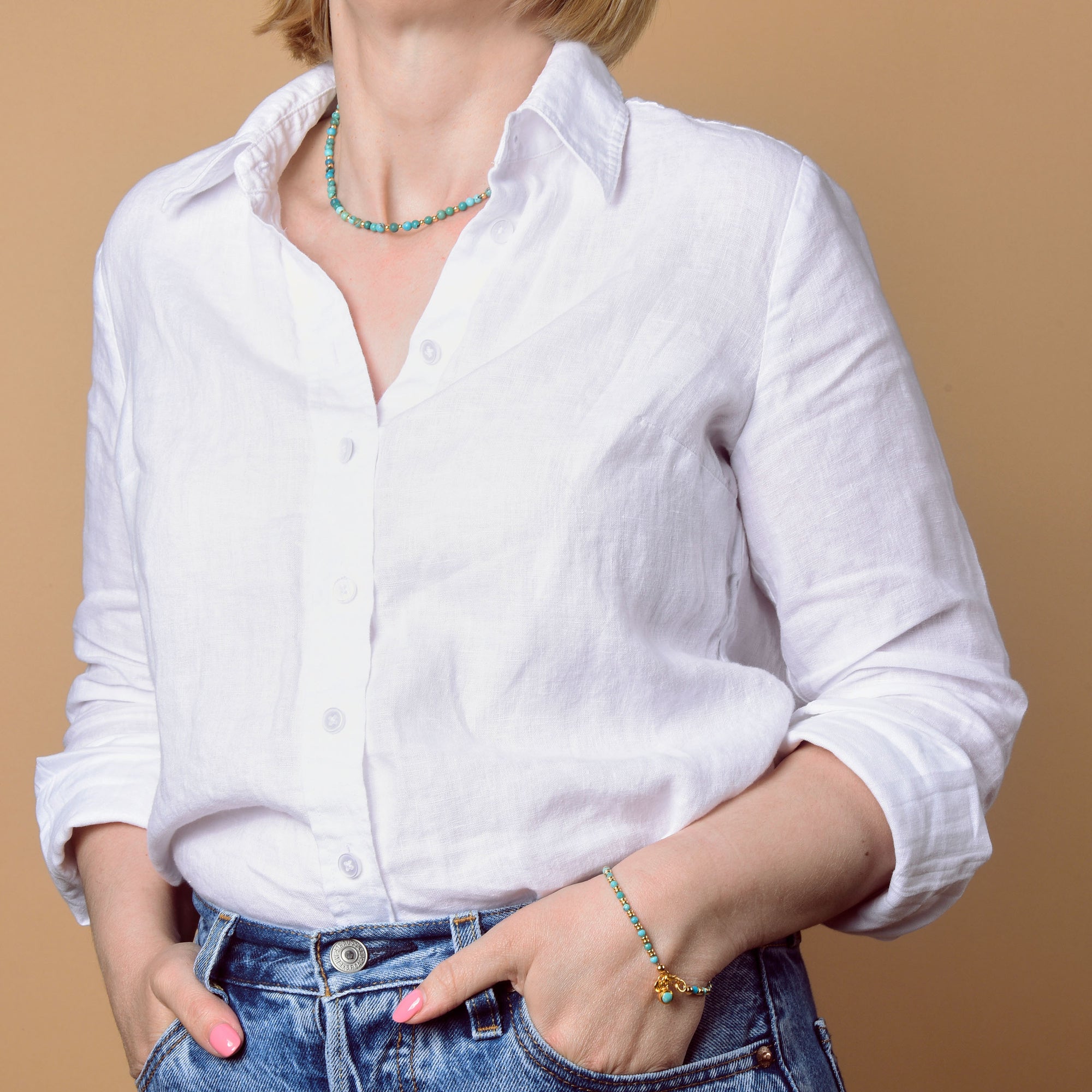 A woman in a white button-up shirt and blue jeans stands with one hand in her pocket, modeling the Jewel Bead Birthstone Bracelet - December against a beige background. Her head is partially out of frame.