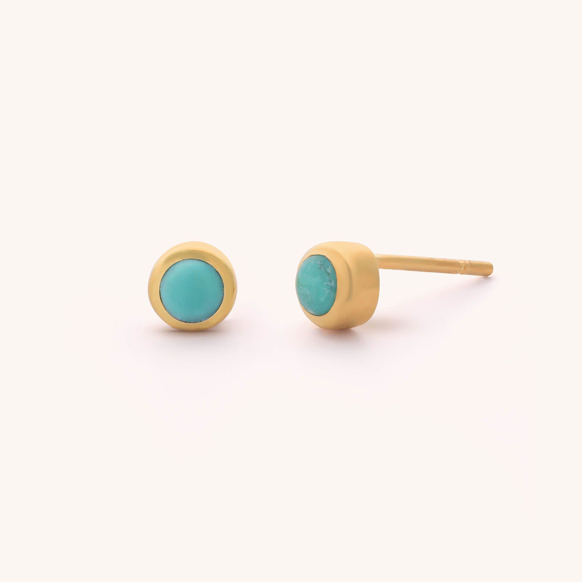 The Mini Gem Dot Birthstone Earrings - December feature elegant gold studs with round turquoise gemstones. One earring displays its vibrant turquoise birthstone from the front, while the other is shown on its side, highlighting the metal post and setting against a plain white background.