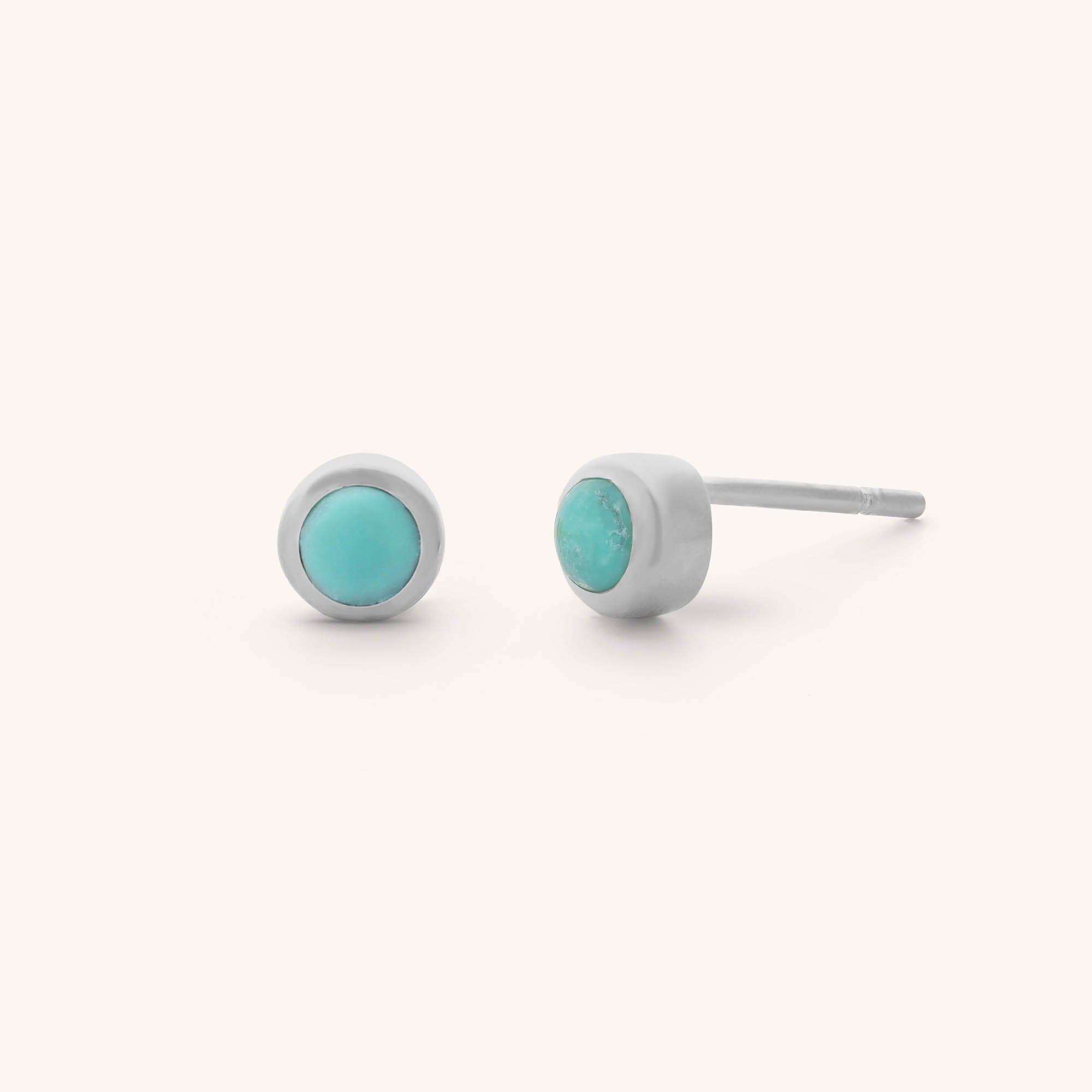 The Mini Gem Dot Birthstone Earrings for December feature two silver studs each with a central round turquoise gemstone on a plain light background.