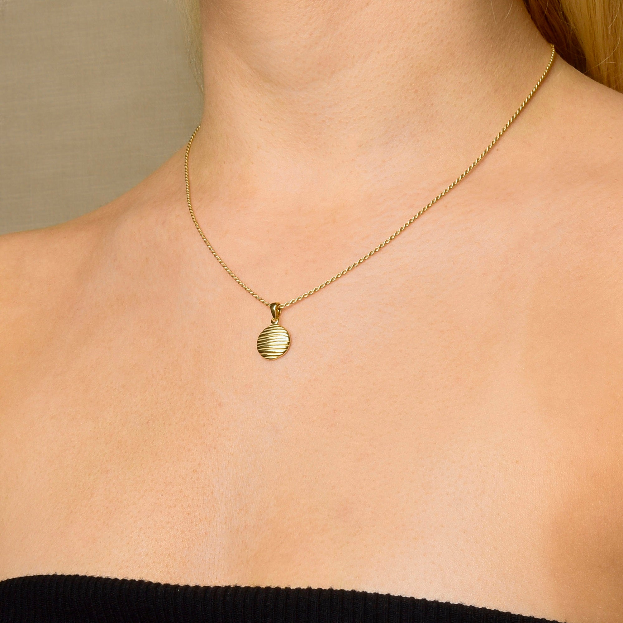 A woman exudes modern elegance wearing the Wave Disc Necklace, featuring a round, ribbed pendant on an adjustable rope chain. Her light skin and black strapless top contrast beautifully with the neutral backdrop.