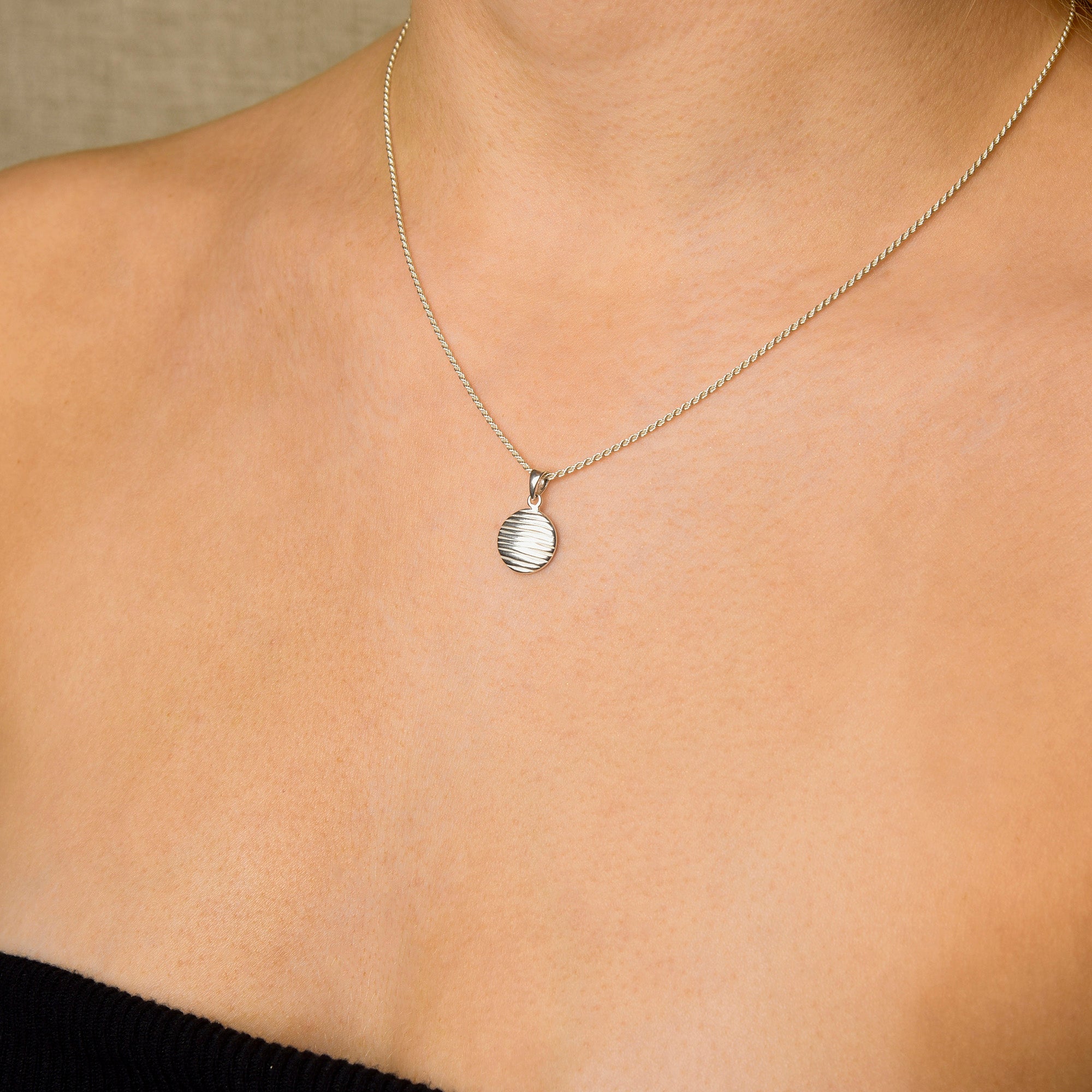 Close-up of a person wearing the Wave Disc Necklace—a round, textured pendant on an adjustable rope chain—styled with a black strapless top for a look of modern elegance against a neutral background.