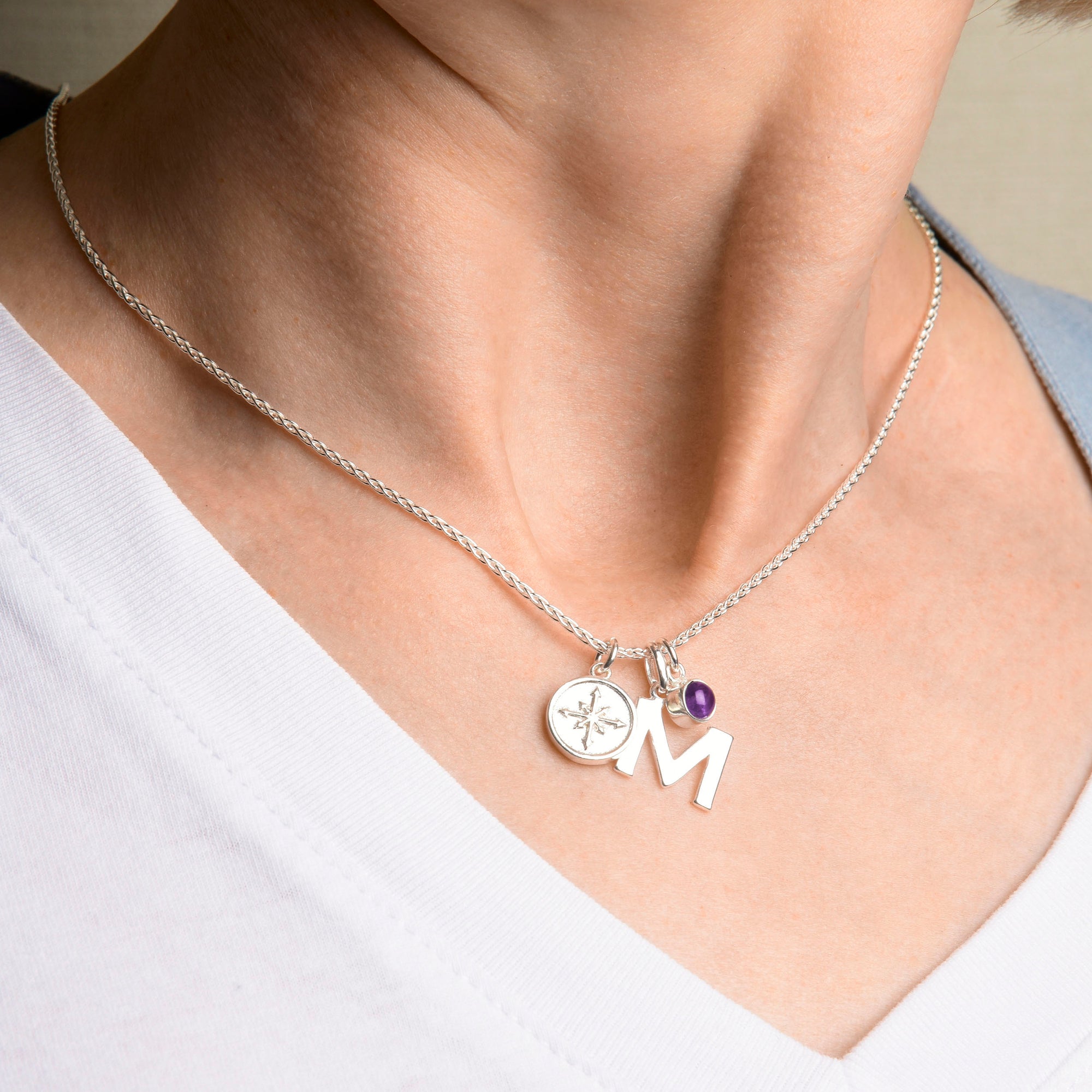 A close-up of a person wearing a white V-neck shirt and a personalised Eco Sterling Silver charm necklace on a Wheat Chain, with a round engraved disc, purple bead, and large silver letter M.