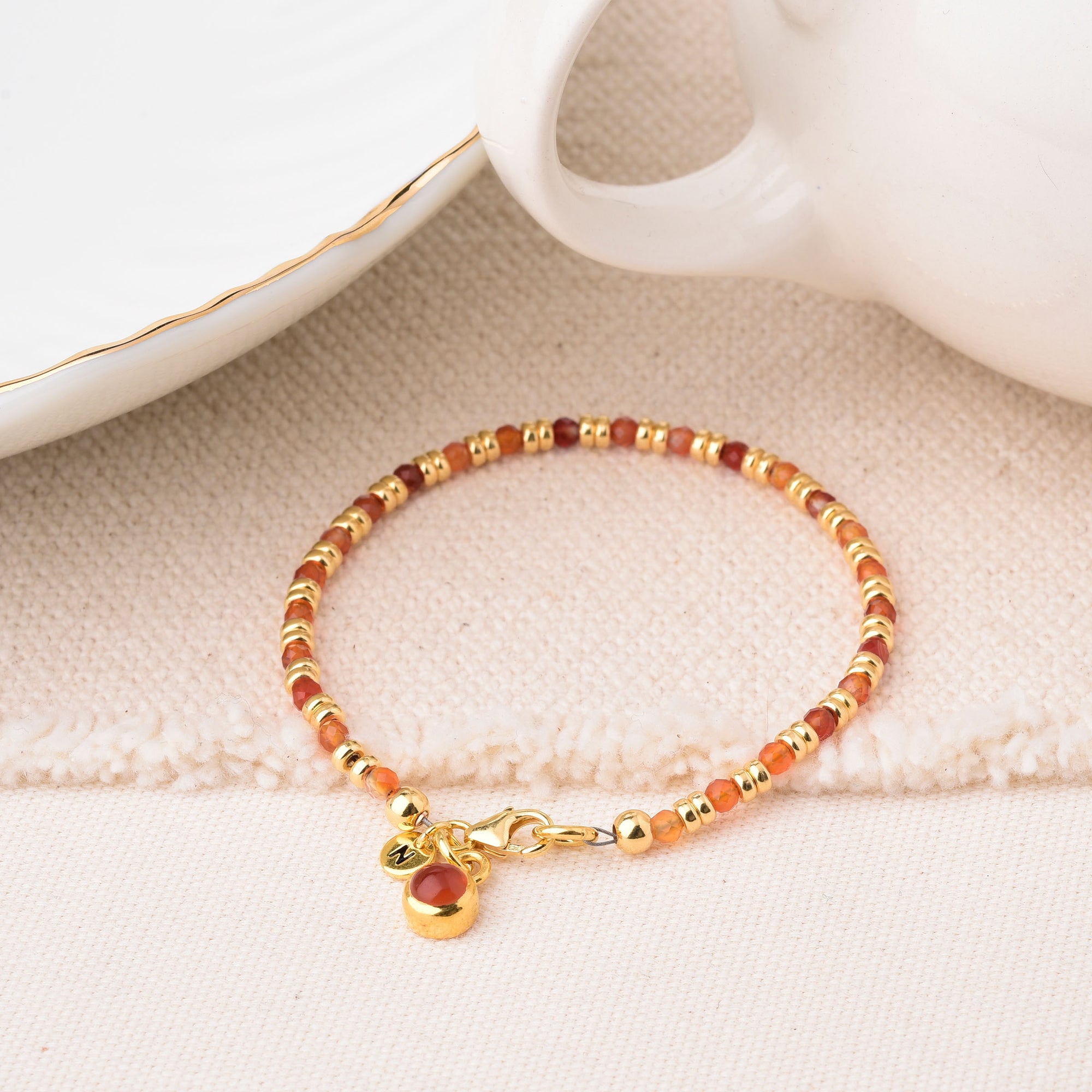 The Linear Birthstone Bracelet - July, a delicate Carnelian piece with gold and reddish-brown beads, gold clasp, and round charm, is displayed on beige textured fabric beside white dishes with gold rims.