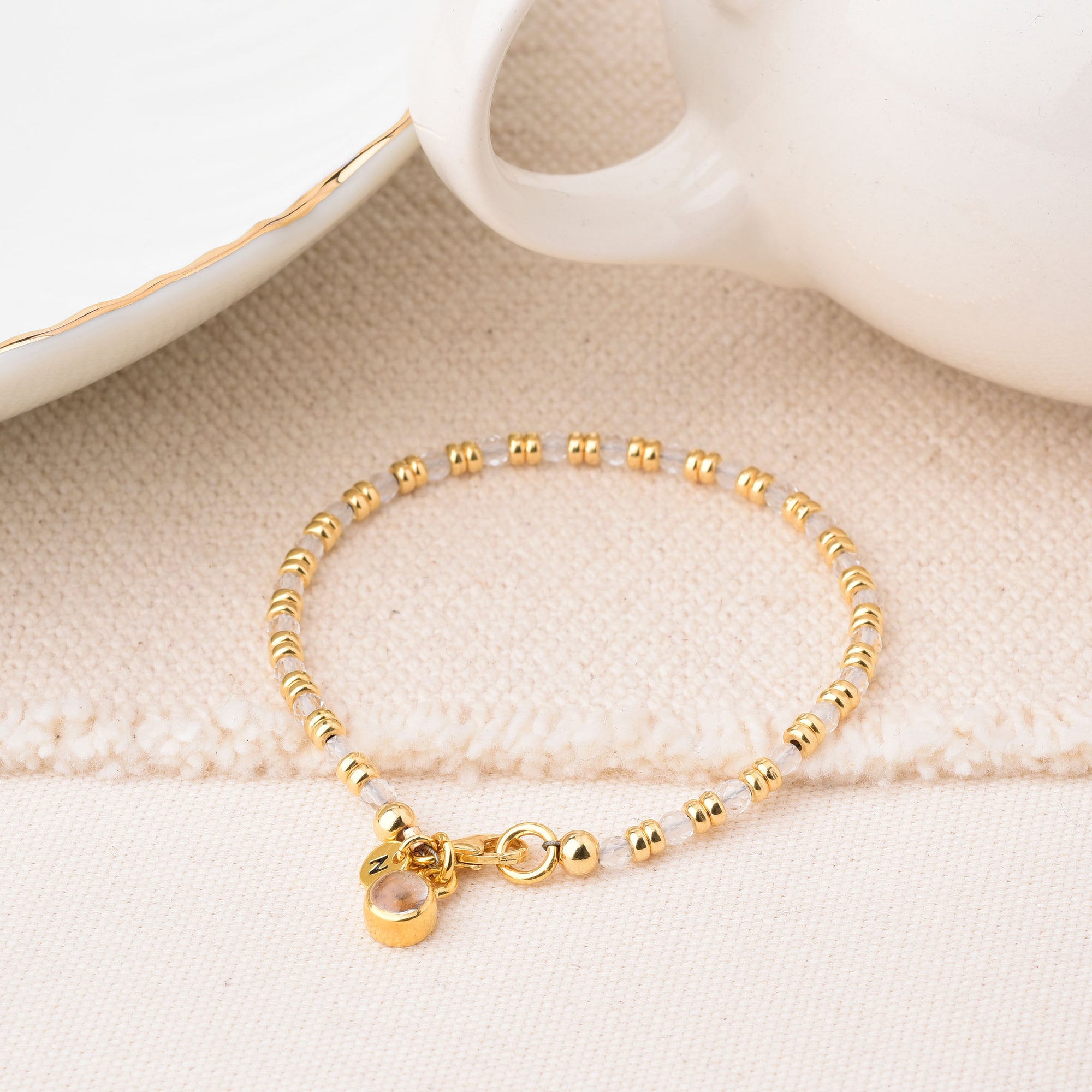 The Linear Birthstone Bracelet is a delicate gold beaded bracelet with a small round charm and gemstone accents, shown on textured fabric next to a white ceramic cup and plate with gold trim.