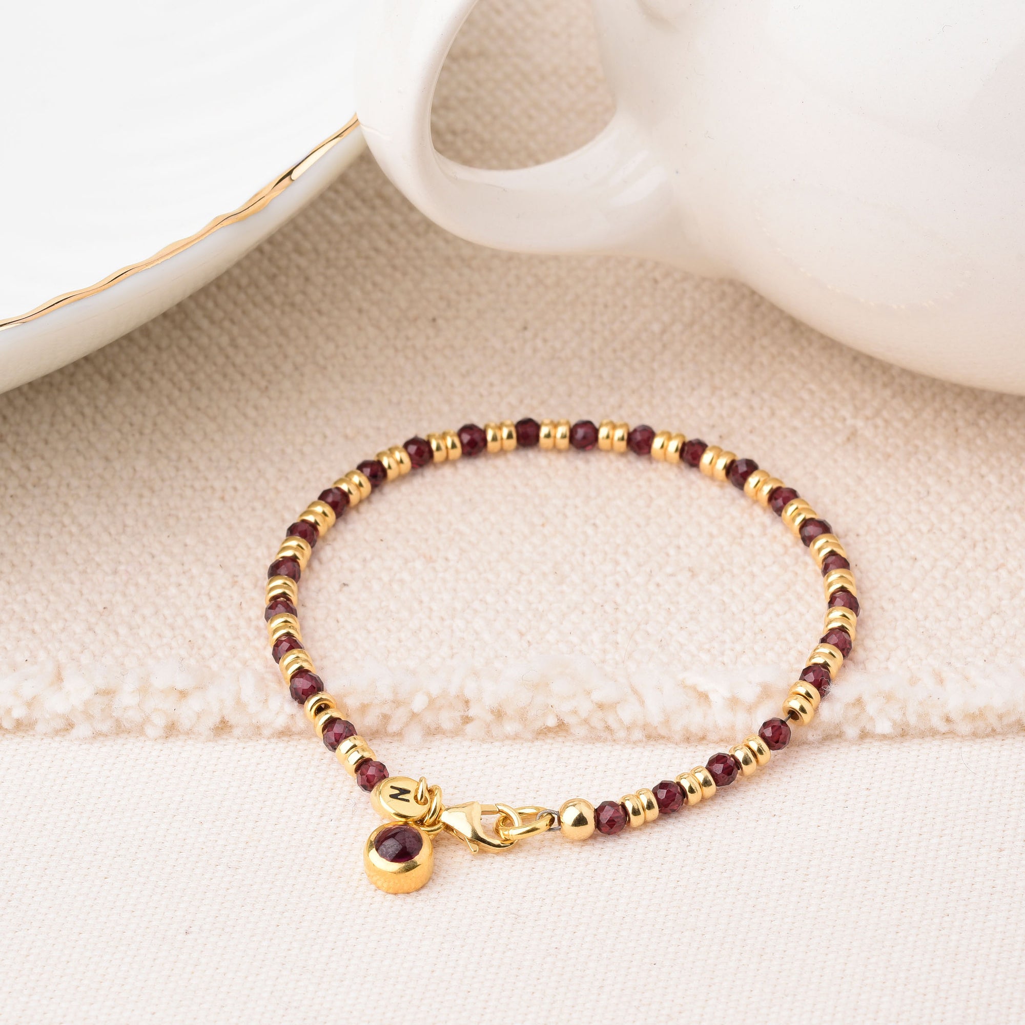 The Linear Birthstone Bracelet - January, with alternating gold and burgundy beads and a small gold charm, is displayed on a light textured surface beside a white teacup and matching saucer.