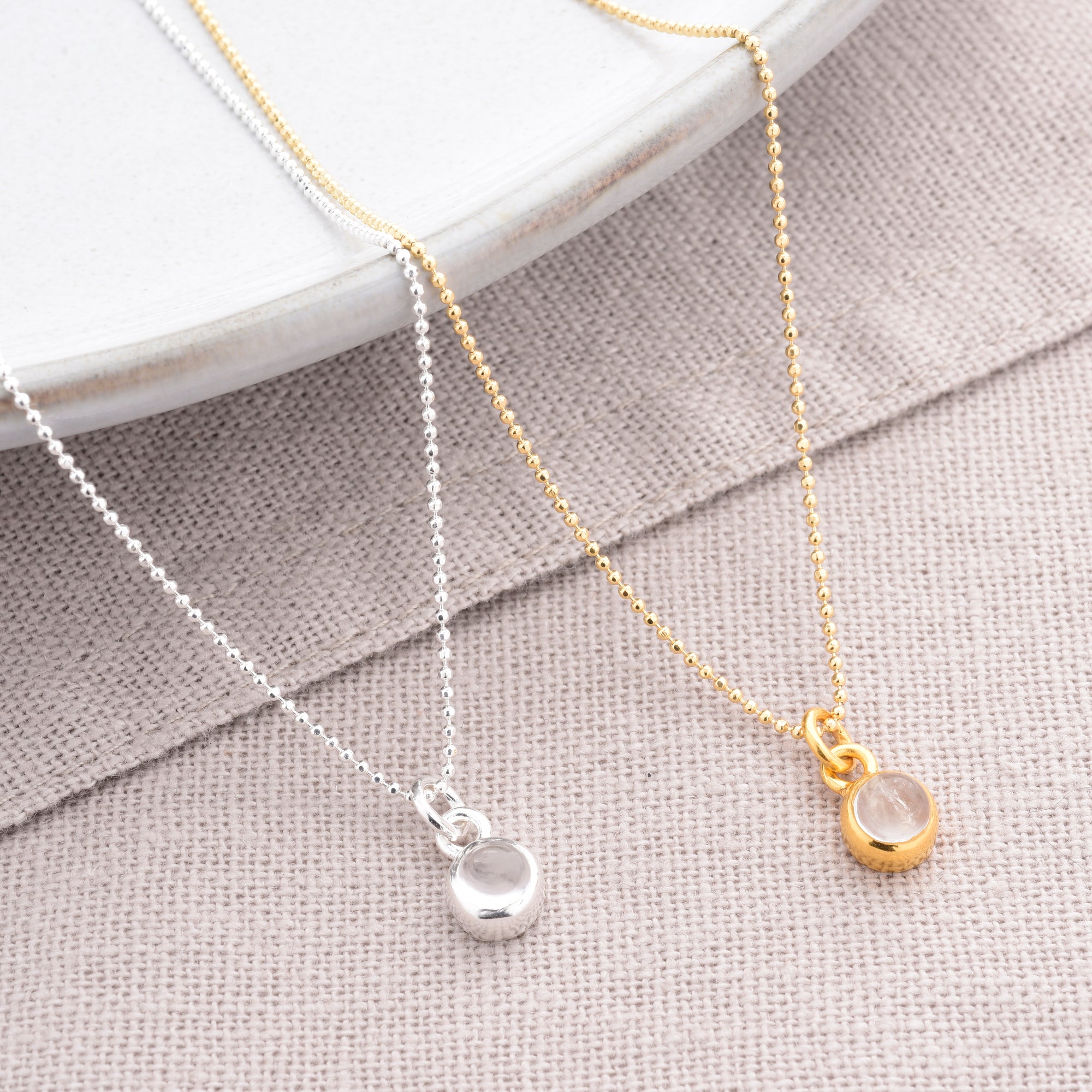 Two Gem Dot Birthstone Necklace April pieces rest on beige fabric—one with a gold chain and bezel-set round pendant, the other with an eco-silver beaded chain. Both feature a clear April birthstone. A white ceramic plate is at the top.