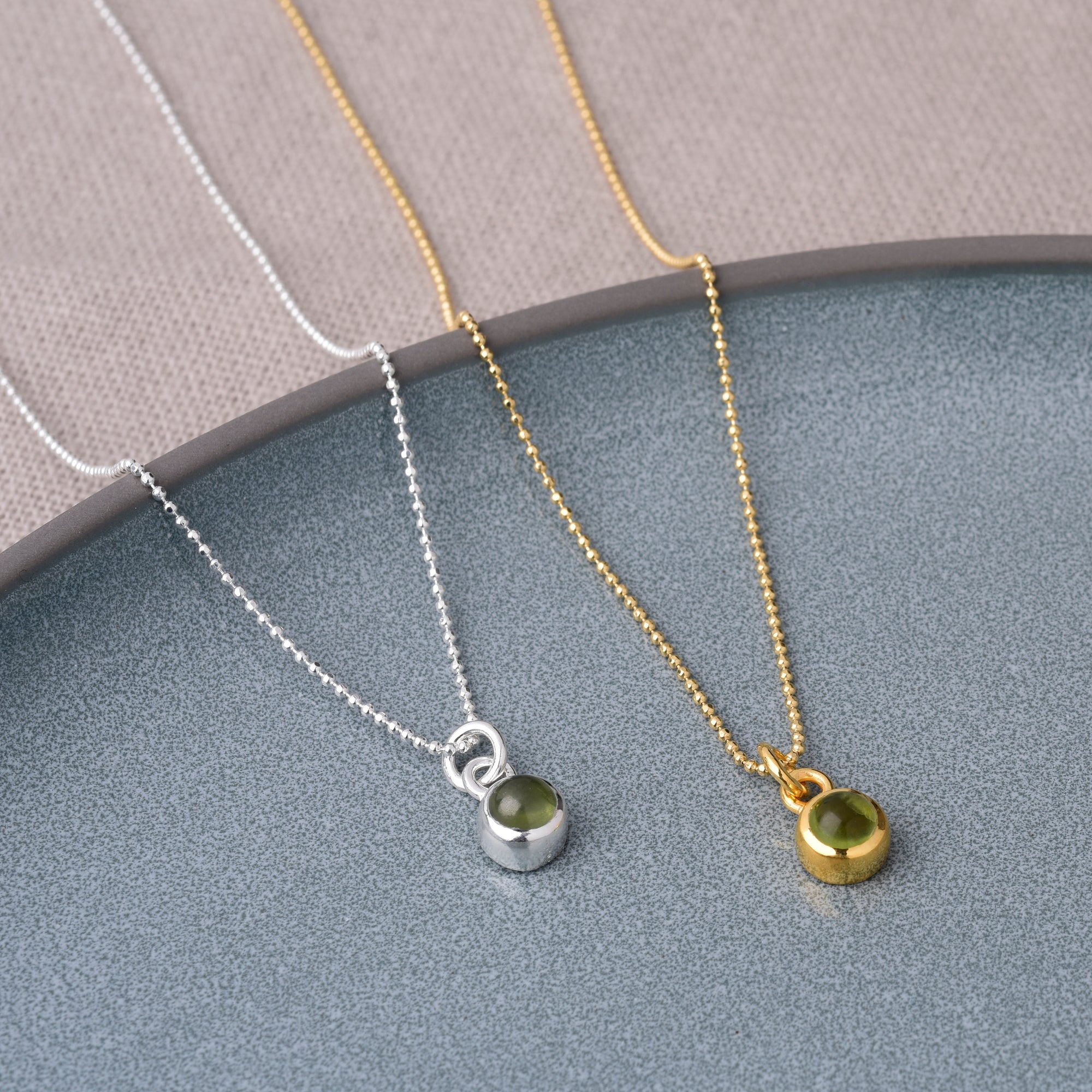 Two Gem Dot Birthstone Necklace August pieces, each with a round Peridot pendant—one in silver and one in gold—are displayed side by side on a gray plate against a beige fabric background.