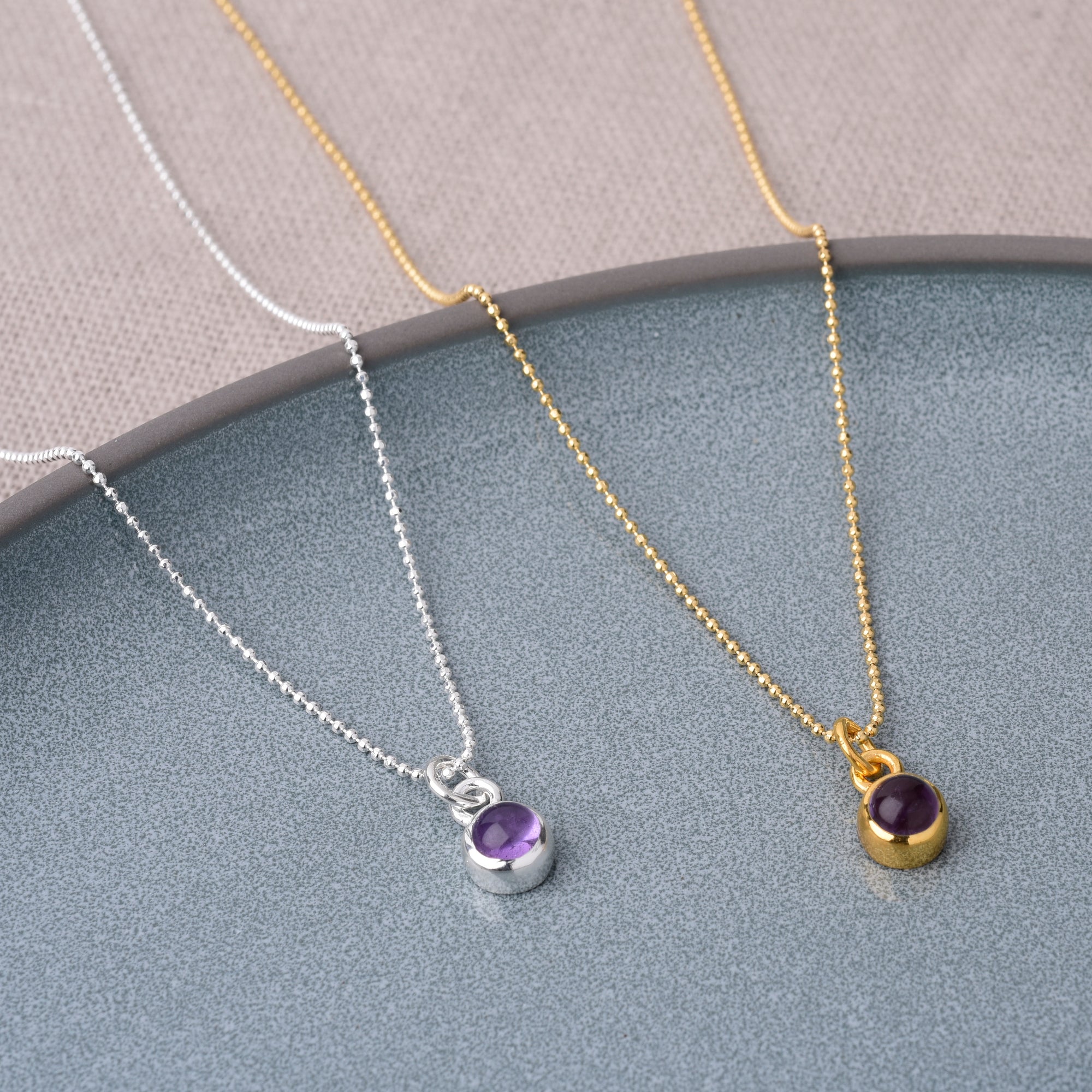 Two Gem Dot Birthstone Necklace February pieces displayed on a grey plate: one with a silver chain and purple Amethyst pendant, one with a gold chain and matching pendant. Both highlight amethysts calming effect in elegant settings.