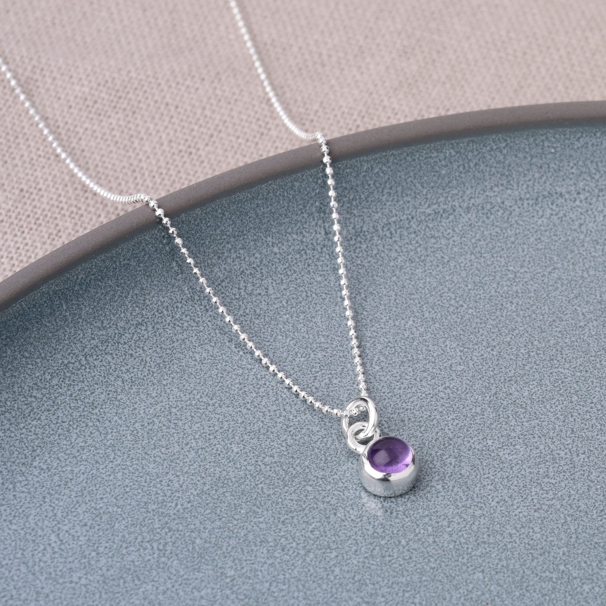 The Gem Dot Birthstone Necklace February, featuring a delicate silver chain with a small round amethyst pendant, is displayed on a gray textured surface beside light beige fabric.