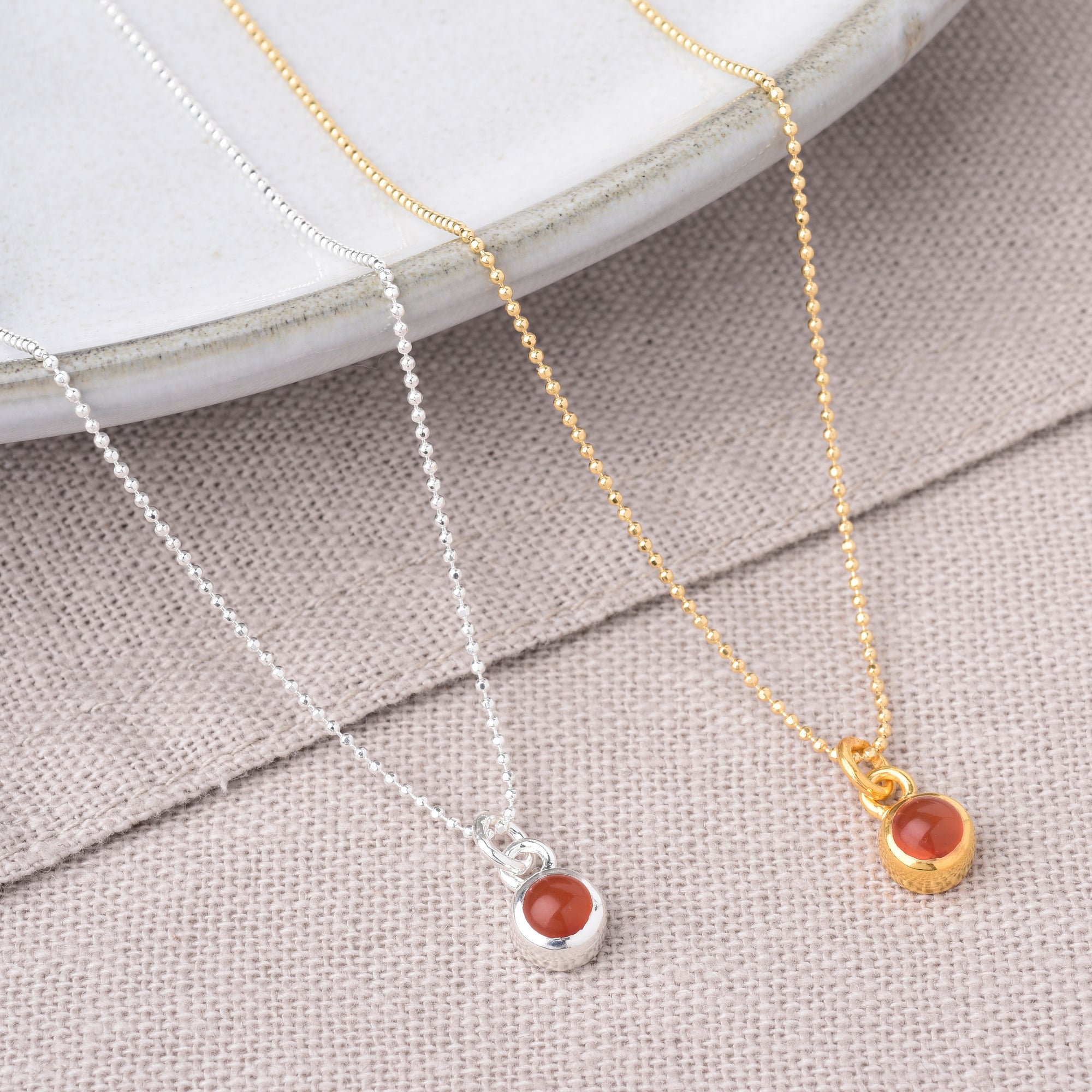 Two Gem Dot Birthstone Necklace July pieces—one in silver, one in gold—each with a small round Carnelian pendant, are displayed on beige textured fabric beside a white ceramic plate.