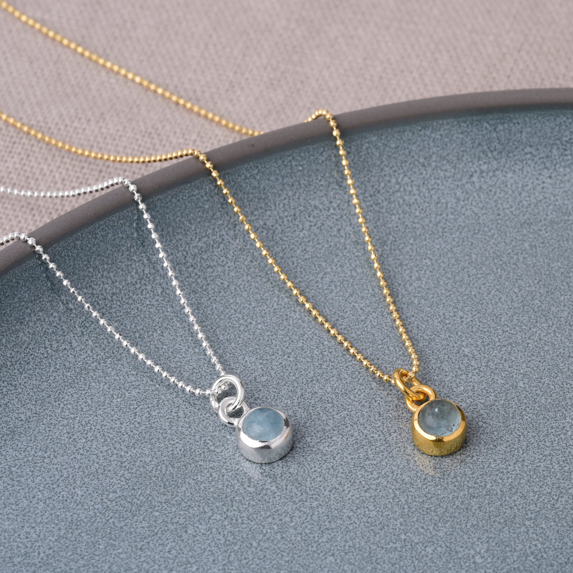 Two Gem Dot Birthstone Necklace March pieces, one silver and one gold, each with a small round aquamarine pendant, are displayed on a textured gray surface.