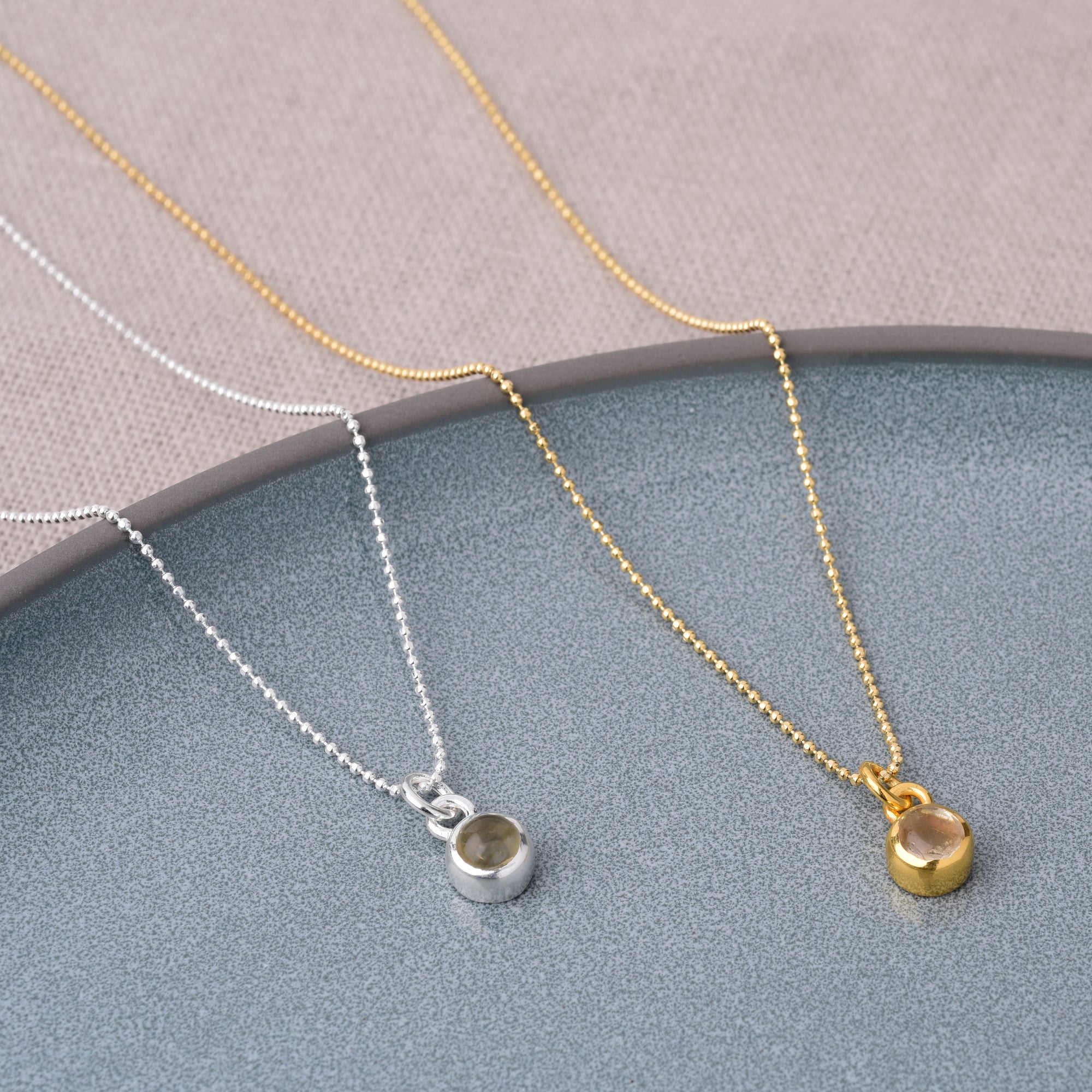 Two Gem Dot Birthstone Necklace November pieces—one radiant Citrine, one classic silver—are displayed on a textured gray surface with a beige fabric background. The luminous November birthstone pendants rest side by side.